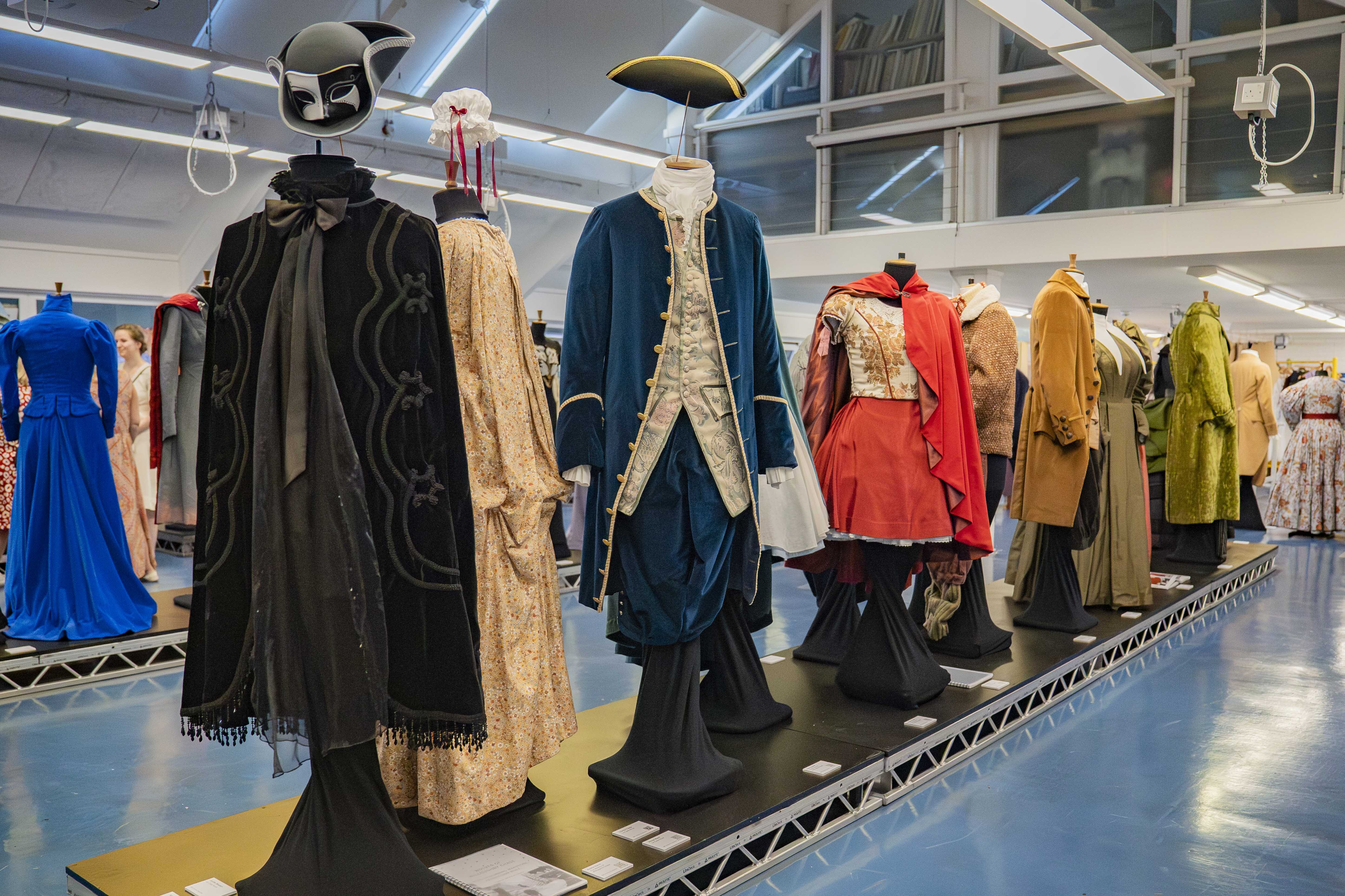 Museum display of historical costumes on mannequins including black cape, blue coat, red military jacket, and colourful period garments.