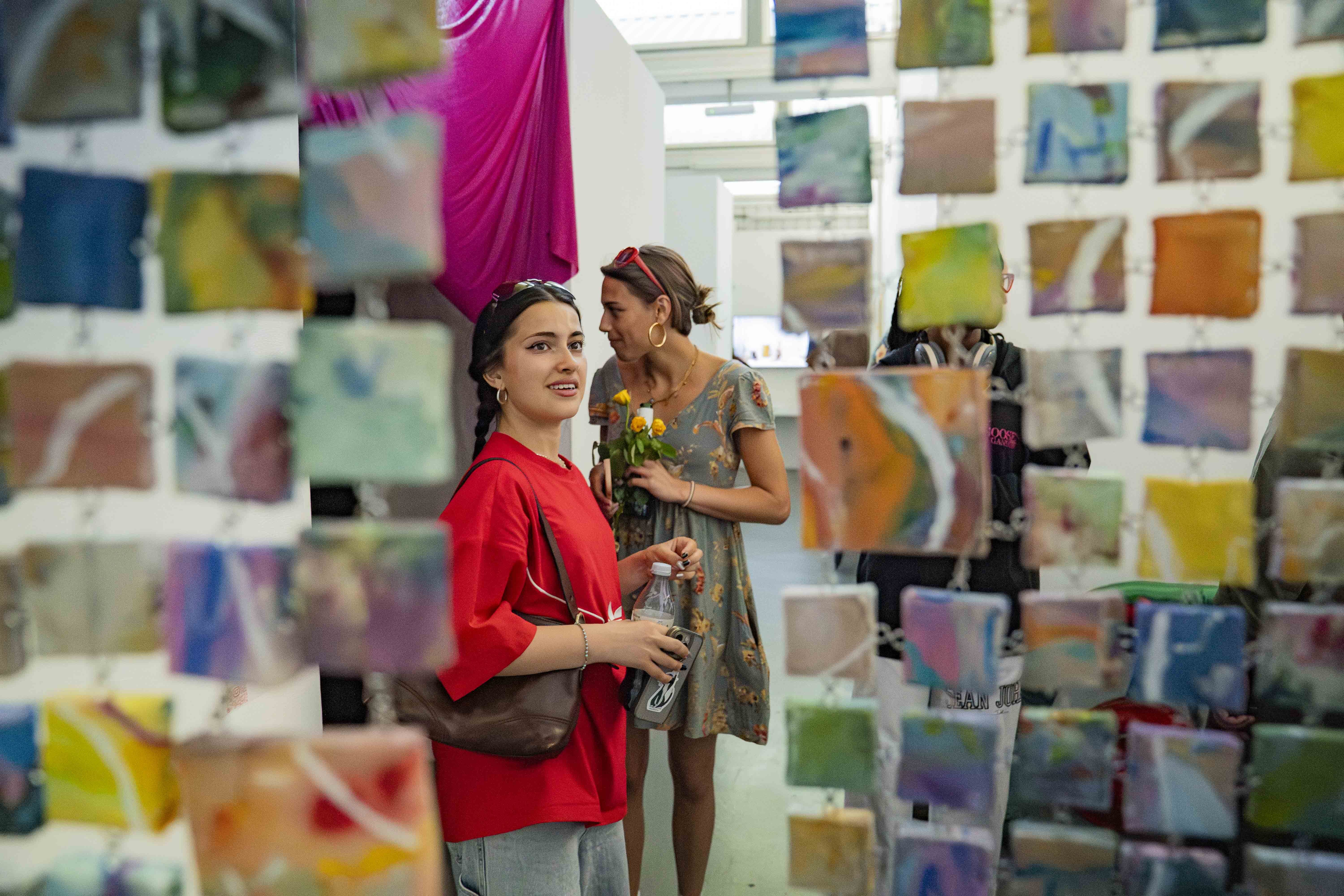 Person in red top stands in art gallery with colourful mini suspended paintings, second person visible in background near camera equipment.
