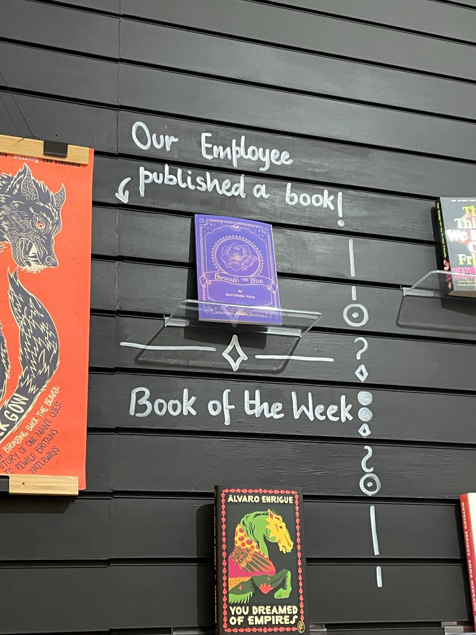 A black wall with Karishma's book 'Beneath the Blue' surrounded by written text: 'Our Employee published a book! Book of the week'.