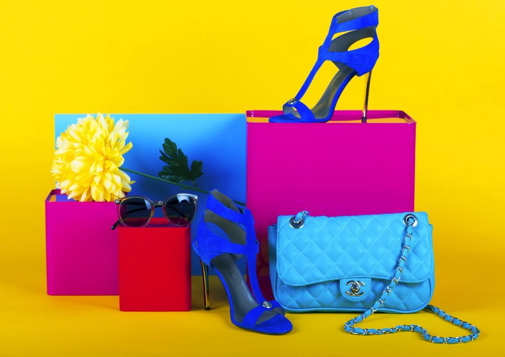 A flower and a pair of sunglasses rest on a pair of boxes. A stiletto heel shoe and a handbag are sitting in front of another box. The other stiletto heel shoe is on top of one of the boxes.