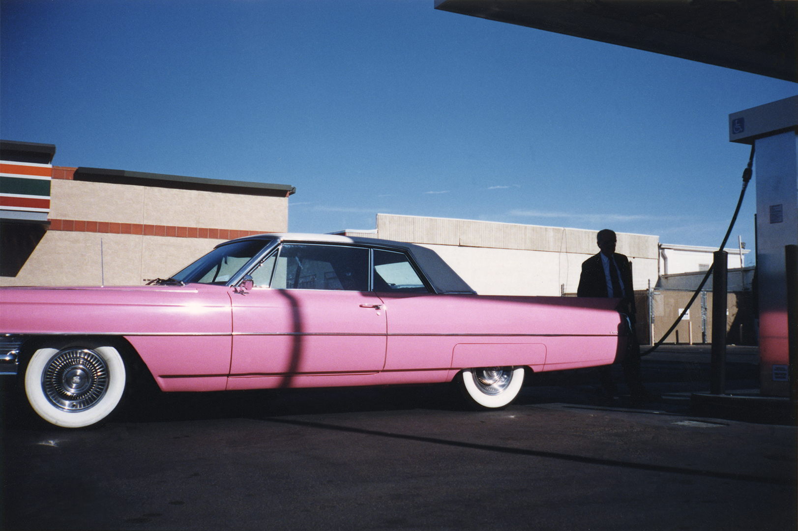 Pink vintage car with whitewall tyres parked beside modernist building under bright blue sky, person in dark clothing walking nearby.