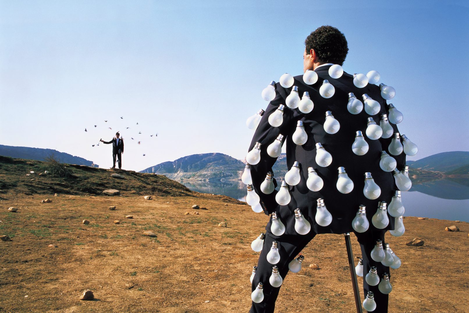 Person in black costume covered with white circular mirrors stands in desert landscape, facing distant figure with arms raised near lake and mountains.