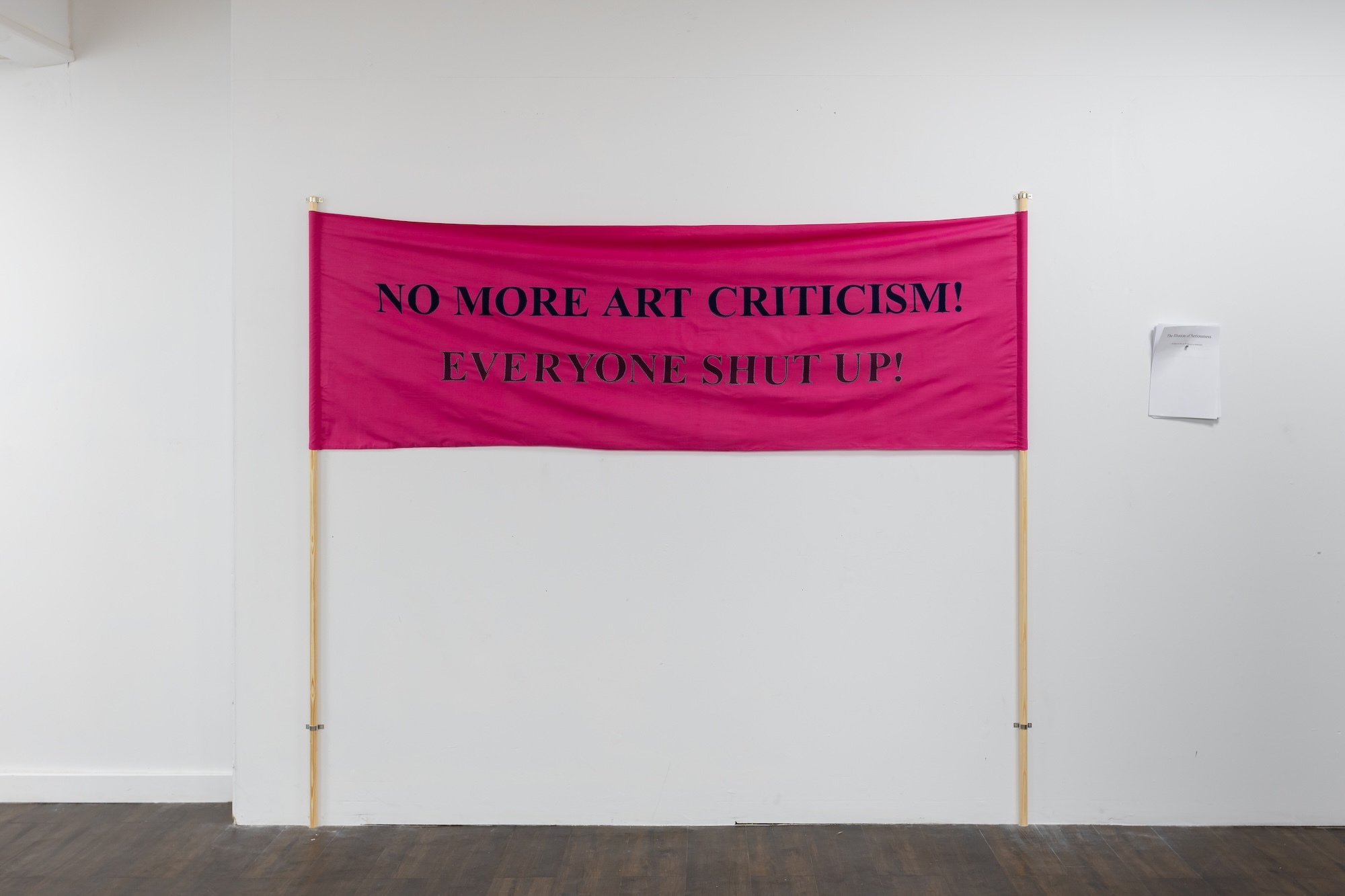 Pink fabric banner with black text "NO MORE ART CRITICISM! EVERYONE SHUT UP" held between two wooden poles against white wall.