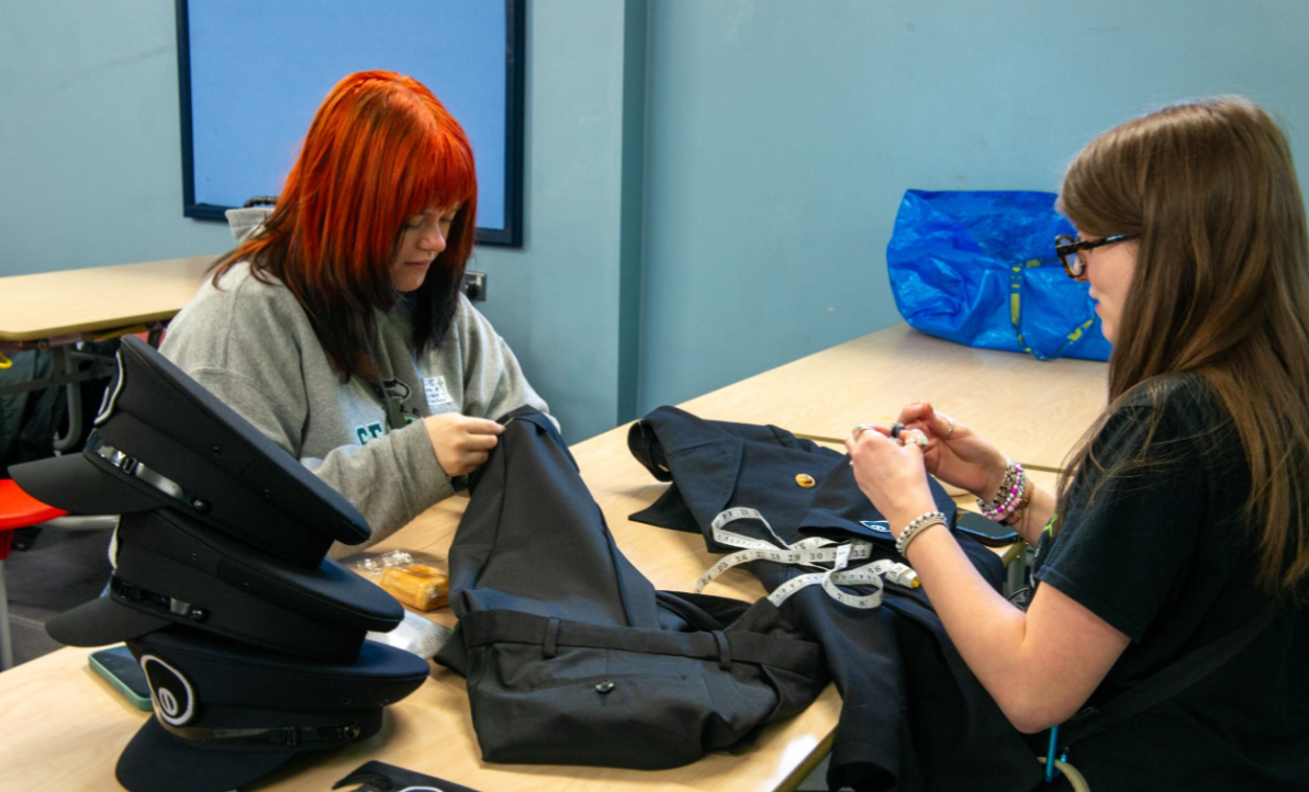 Two BA (Hons) Design for Costume and Performance students sitting either side of a long table. One is making adjustments to a pair of dark-coloured trousers, the other is making adjustments to a long military-style jacket.