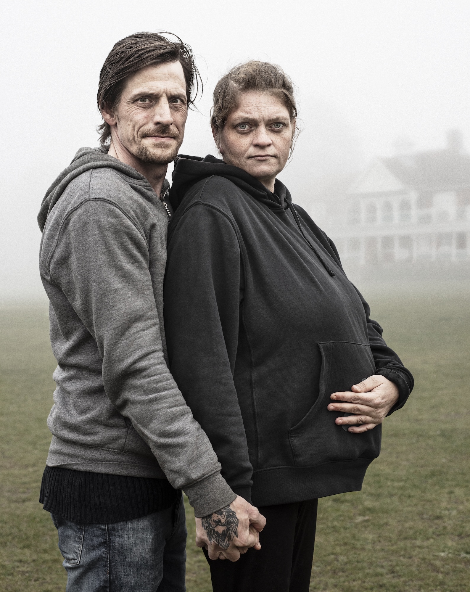 Two people in grey hoodies standing close together outdoors, with blurred countryside and buildings visible in misty background.