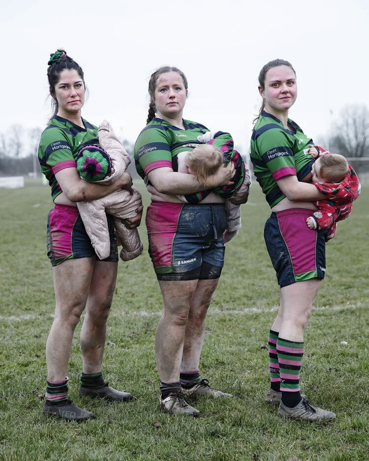 Three women in green and pink rugby kit holding babies, standing on grass field with muddy legs and boots