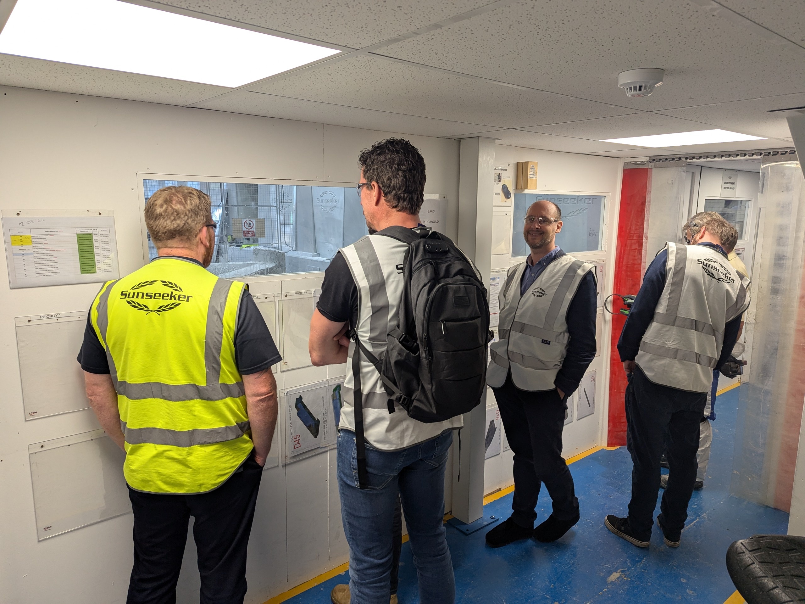 Five men in high-visibility vests and work clothing stand in white corridor with blue floor, viewing wall-mounted displays.