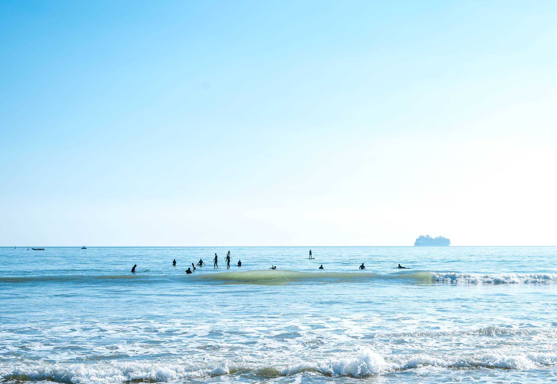 The sea with waves crashing in the foreground and people swimming and paddle-boarding further out.