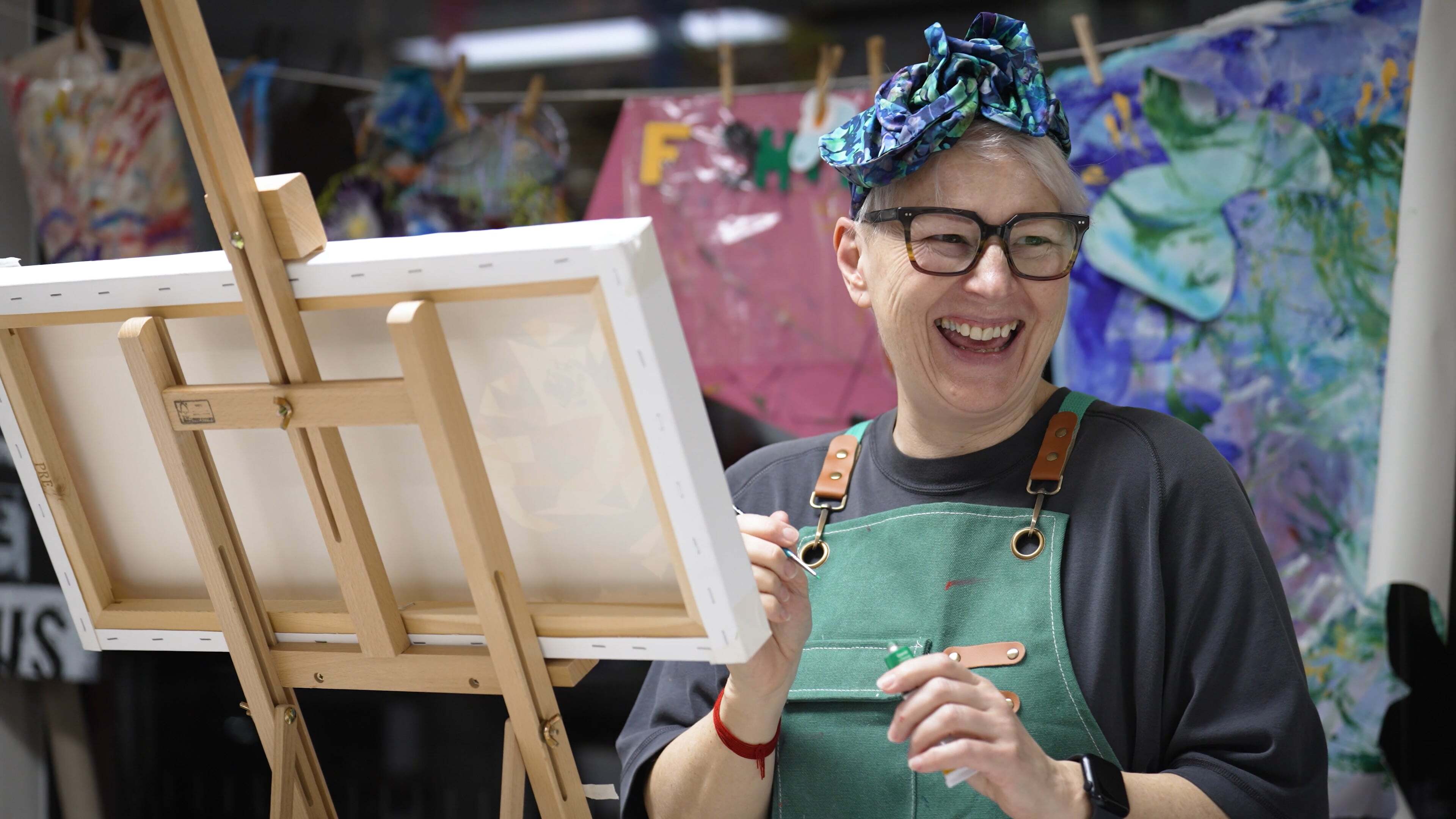 Artist with glasses and colourful headband laughing whilst holding paintbrush, wearing green apron in studio with easel and paintings.