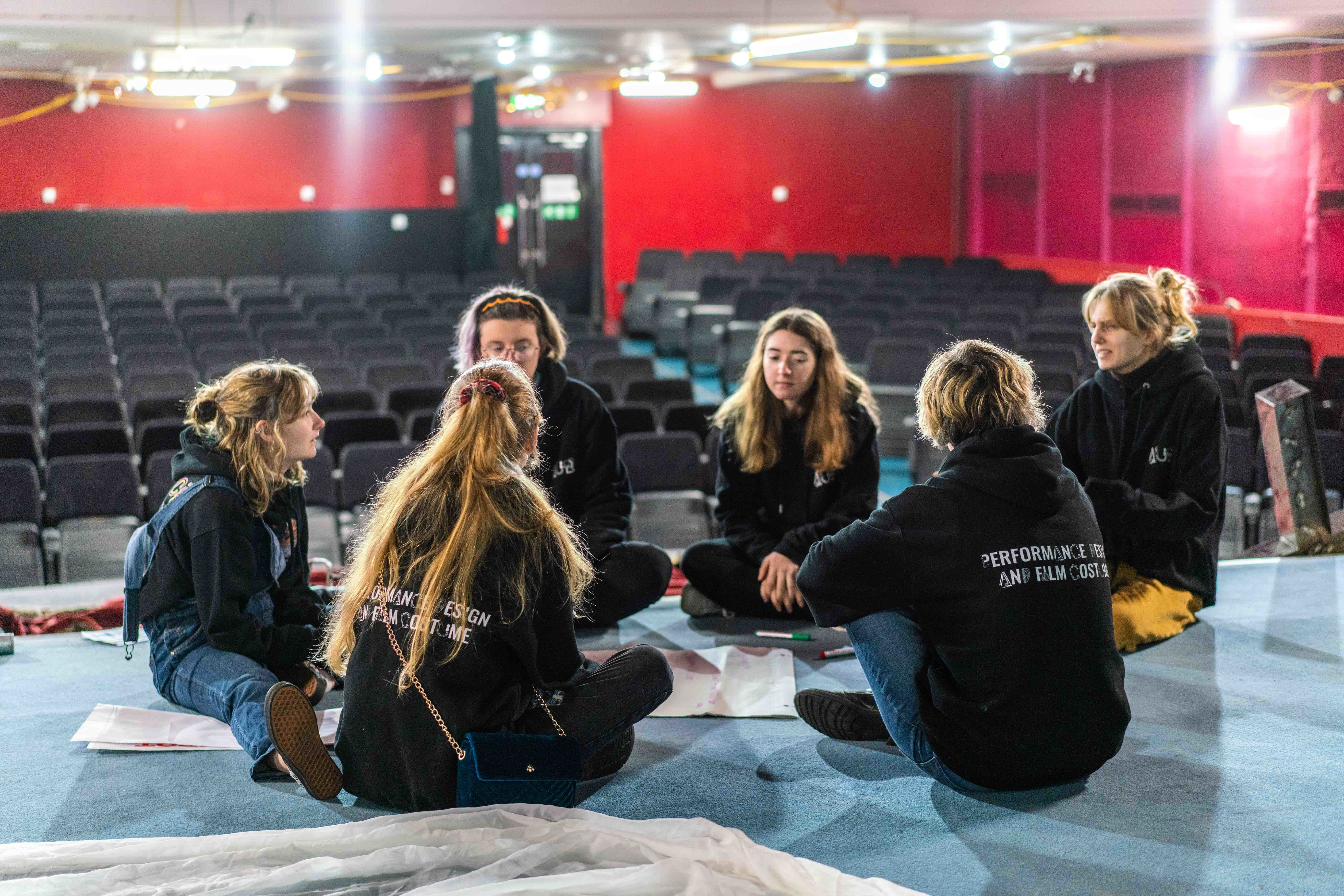 A small group of people sit in a circle on a theatre stage with rows of seats in the background.