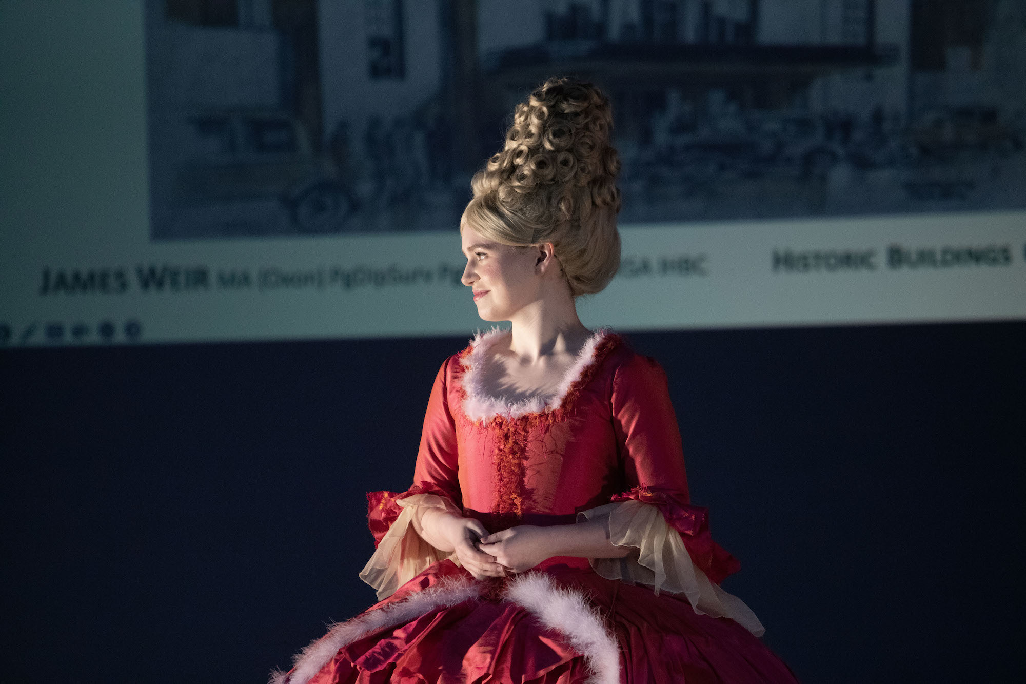 A person in a period costume and wig stands on a stage and looks towards a light on the left.