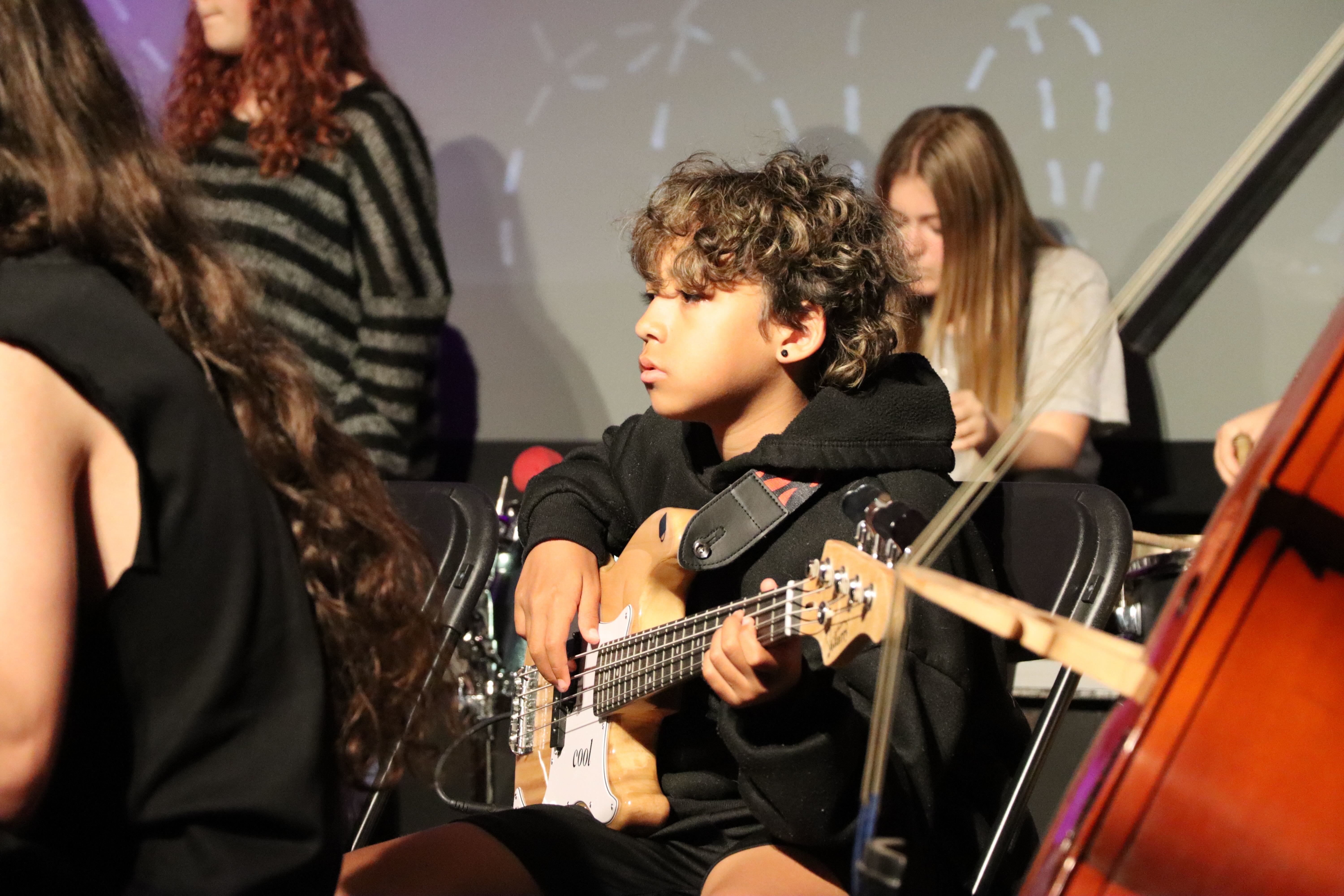 A young person playing an electric guitar, surrounded by other young musicians, on stage at the Palace Court Theatre, Bournemouth.