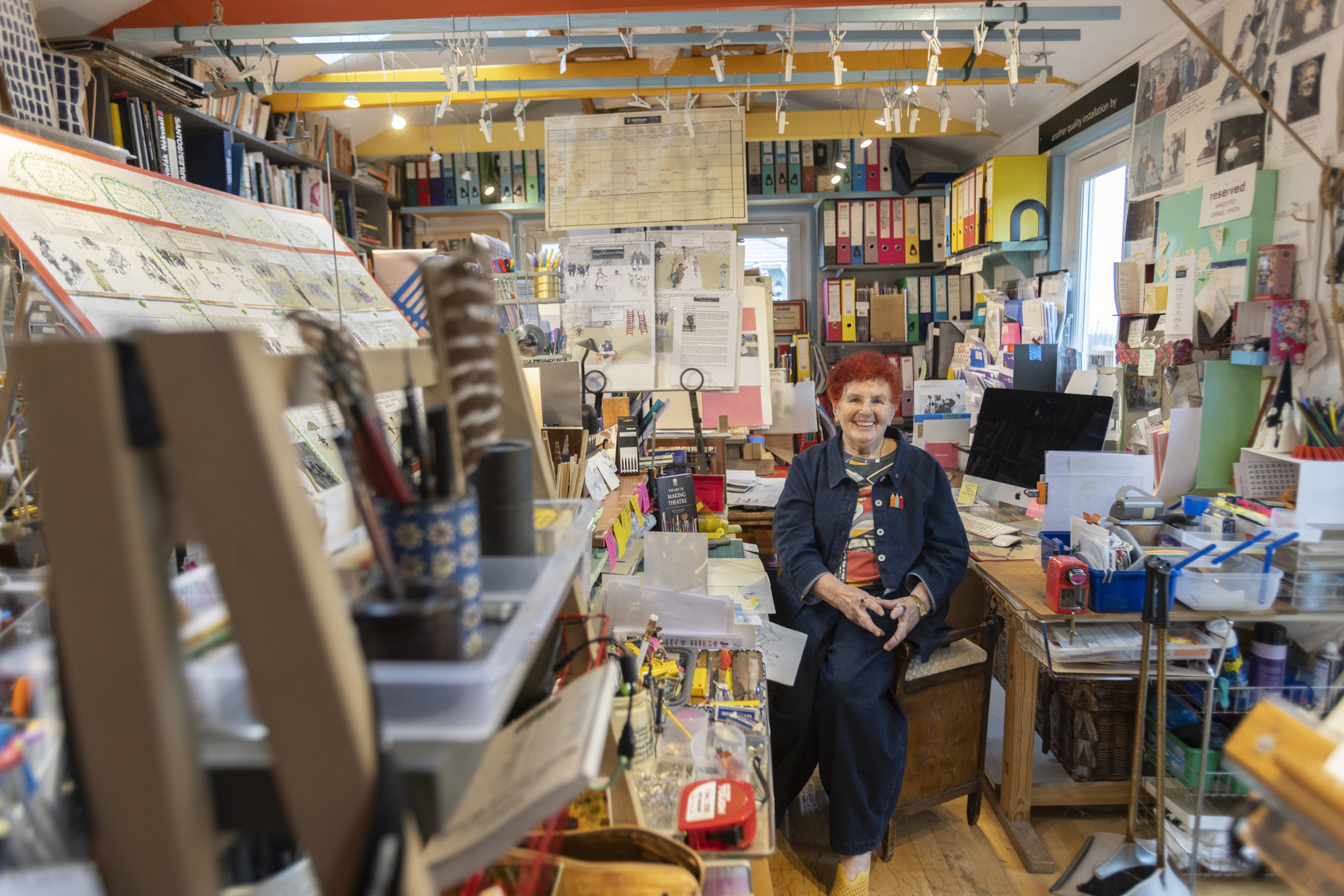 Pamela Howard in a cluttered studio space full of colour and character, with red hair and wearing a blue denim outfit.