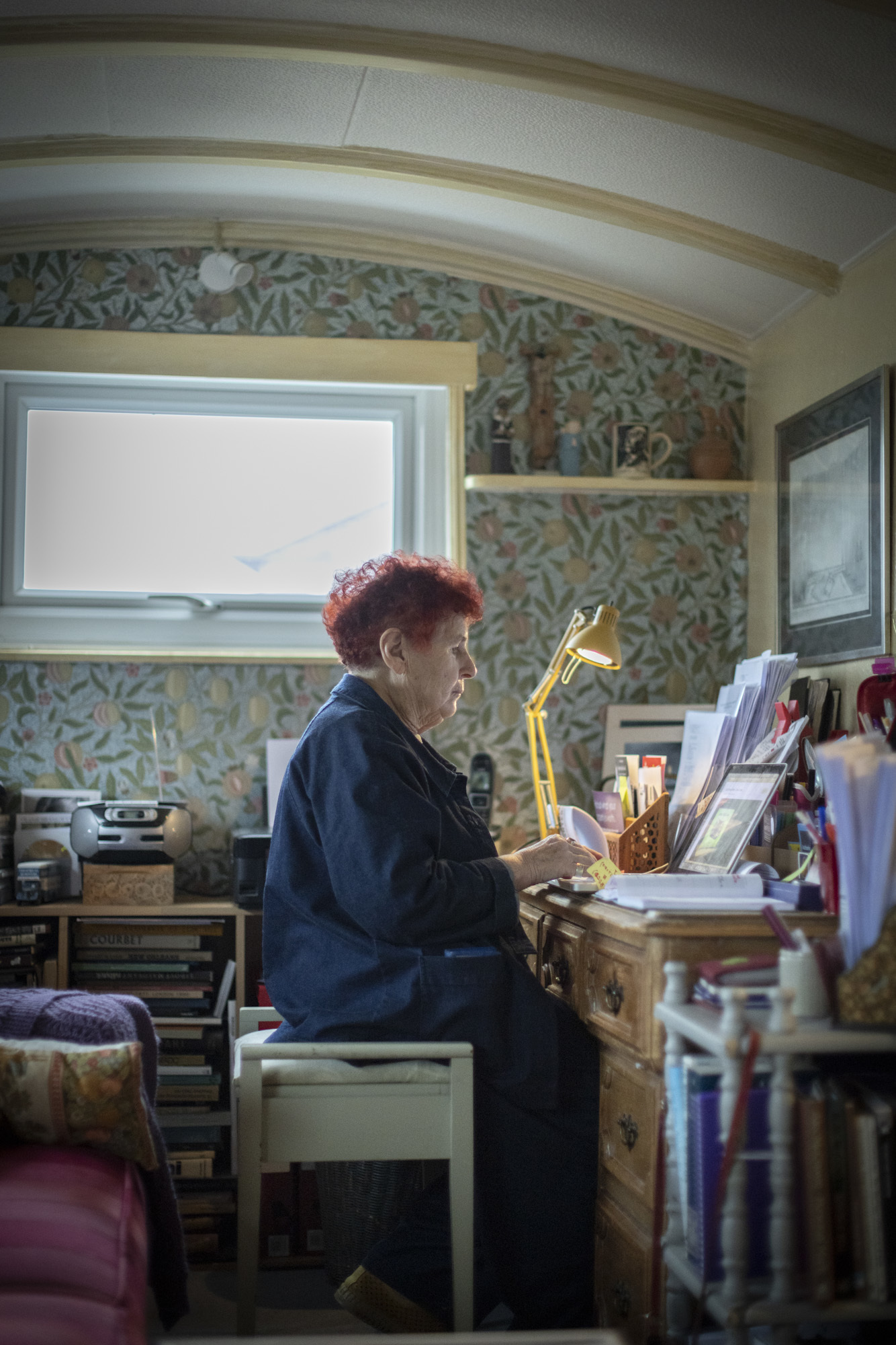 Pamela Howard working in her studio by a yellow lamp on a wooden desk, flower wallpaper in the background.