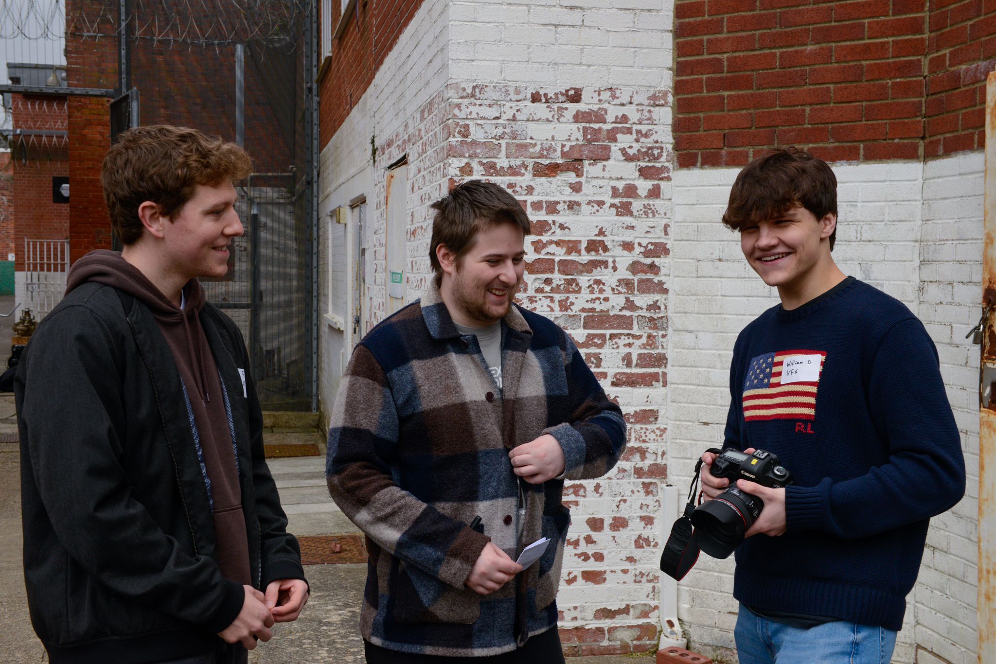Three young men standing outdoors against brick wall, one wearing dark hoodie, one in striped jumper, one in American flag shirt holding camera.