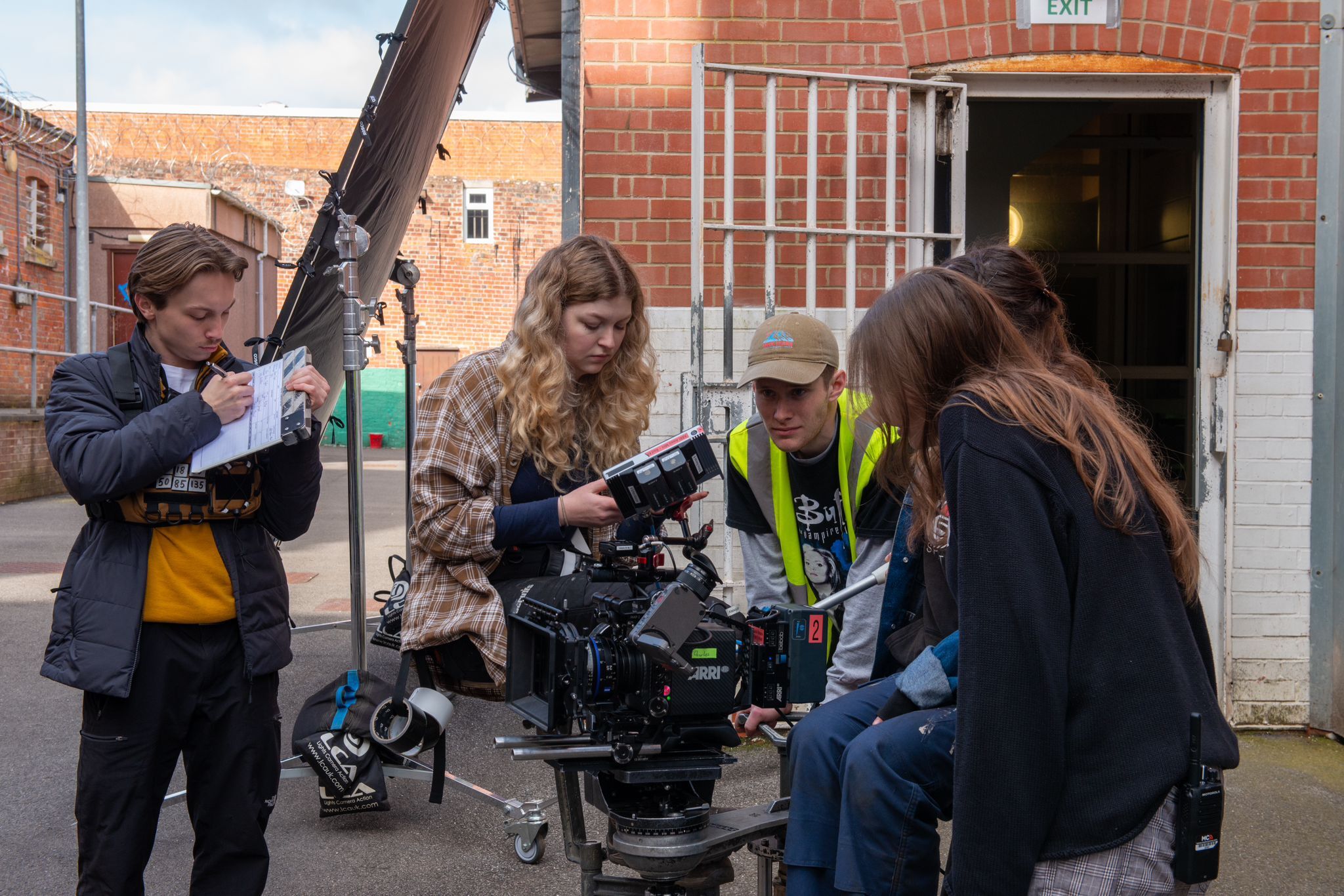 Film crew operating camera equipment on street with brick buildings. Four people gathered around professional camera on tripod.