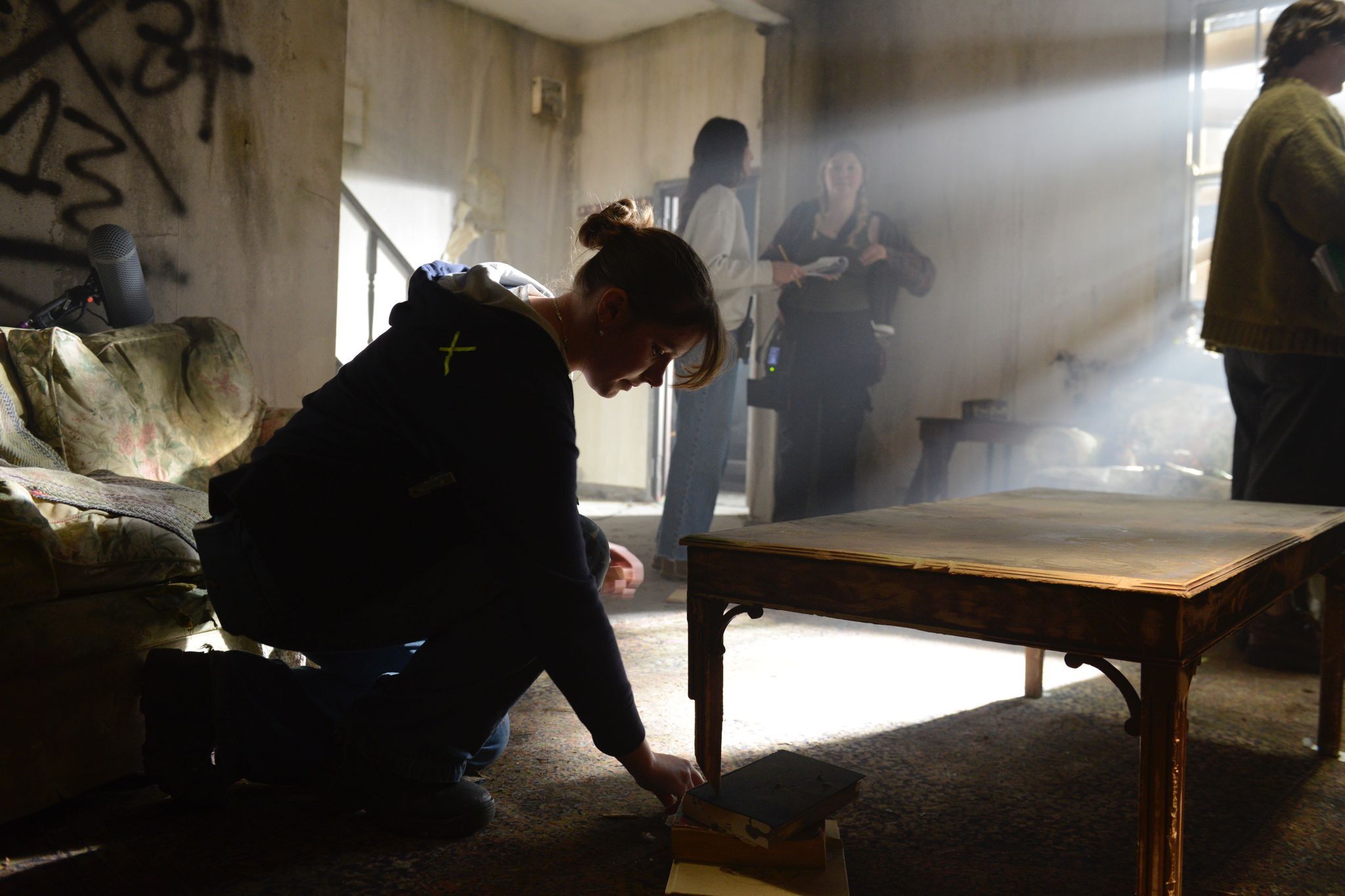 Person kneeling on floor in smoky indoor scene with wooden table, military personnel visible in background, beige walls and window.