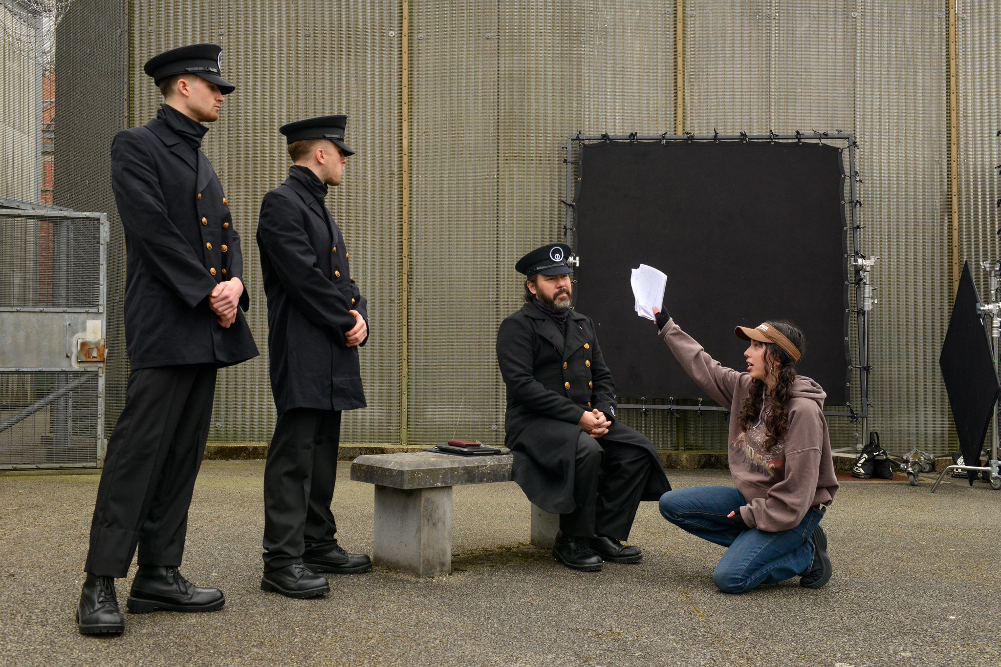 Three police officers in dark uniforms stand and sit near metal building whilst person in brown jacket kneels holding white paper.