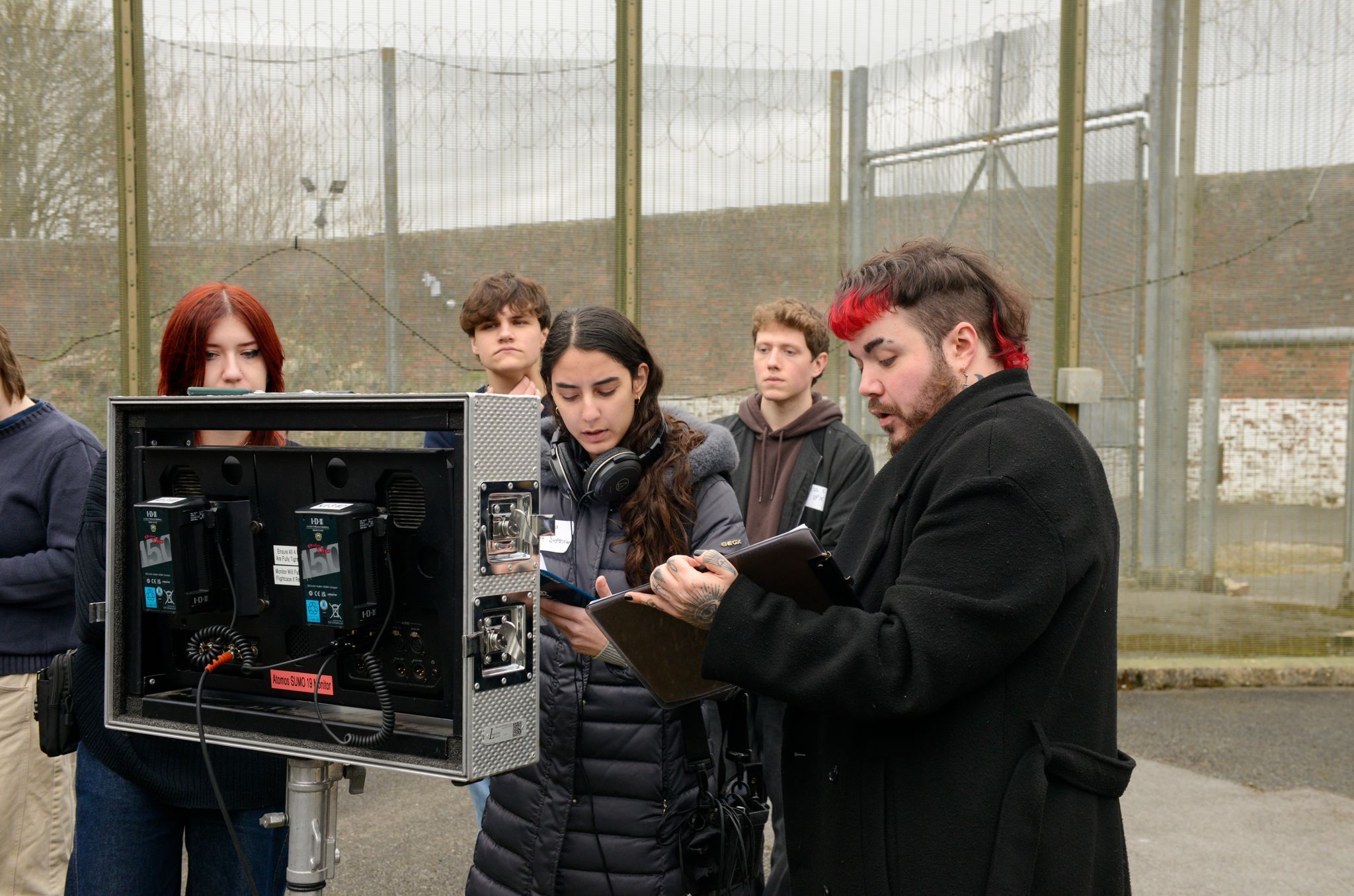 Film crew gathered around monitor on set, with director and actors reviewing footage in industrial glass-walled location.