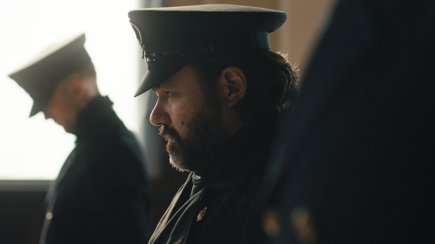 Bearded man in military cap and dark uniform sitting indoors, another uniformed figure visible in background, warm lighting.