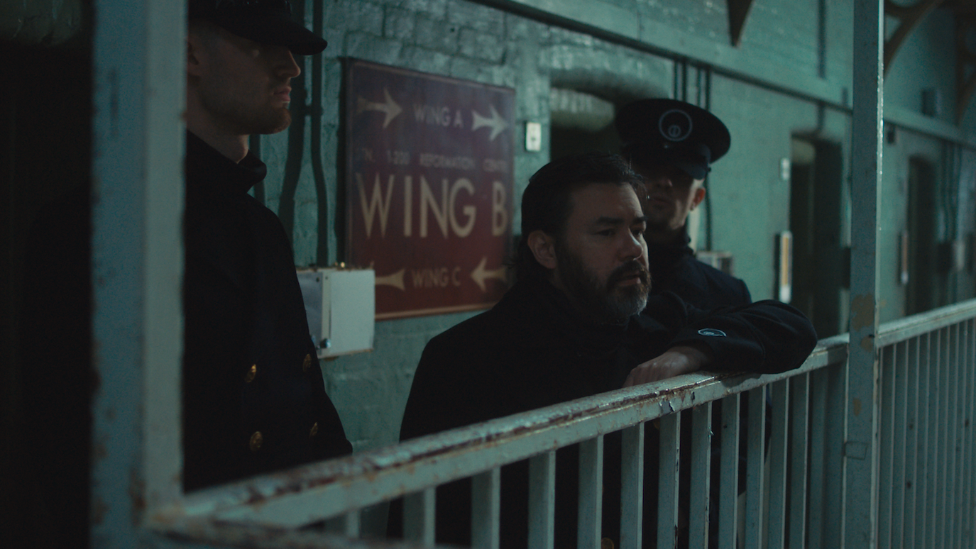 Man in dark coat and cap standing on ship deck with white railings, dimly lit with greenish tones and "WING" sign visible.