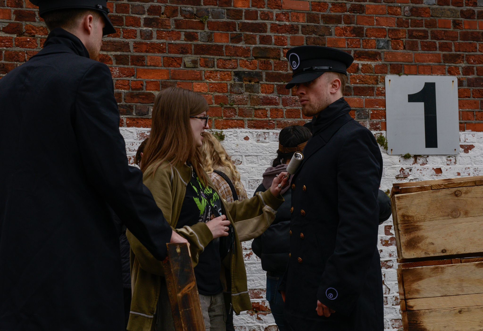 Police officers speaking with woman in green jacket near brick wall with number 1 sign and wooden crates.