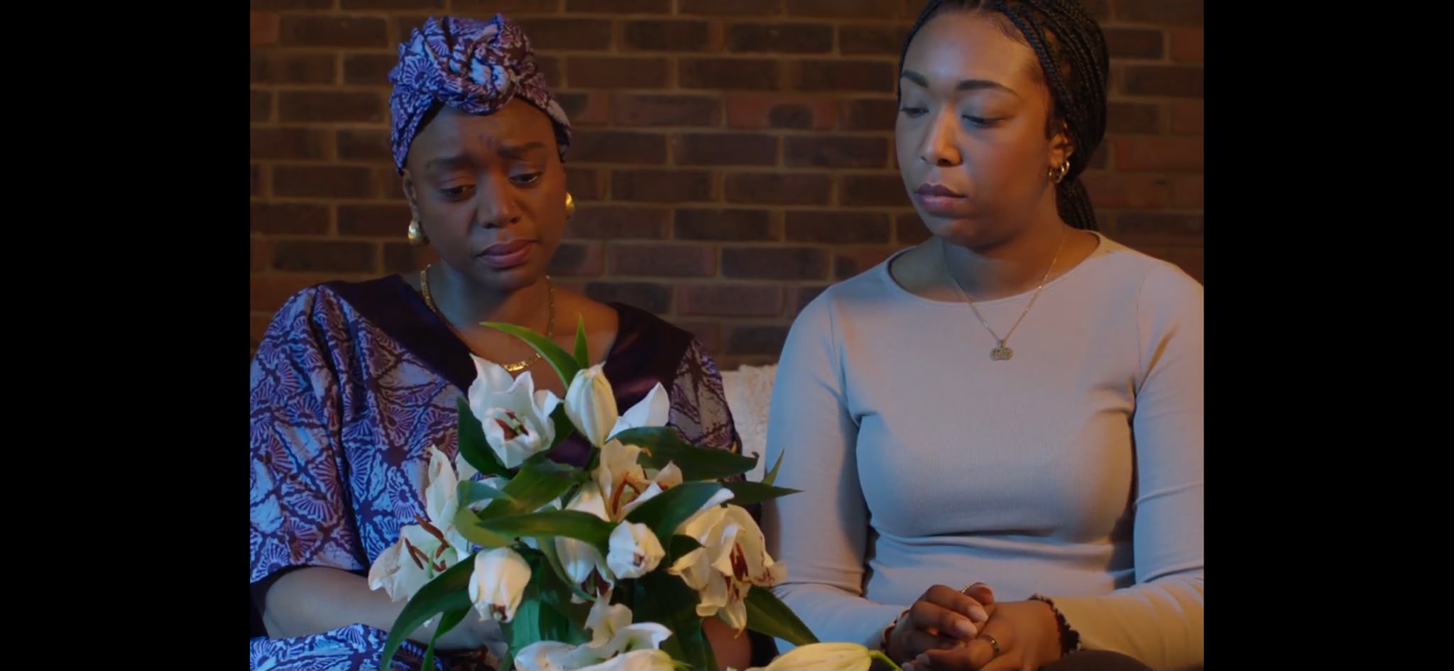 A woman in traditional African attire sits next to a younger woman, both looking down towards a bouquet of white flowers placed in front of them. The background features a brick wall, creating a somber atmosphere.