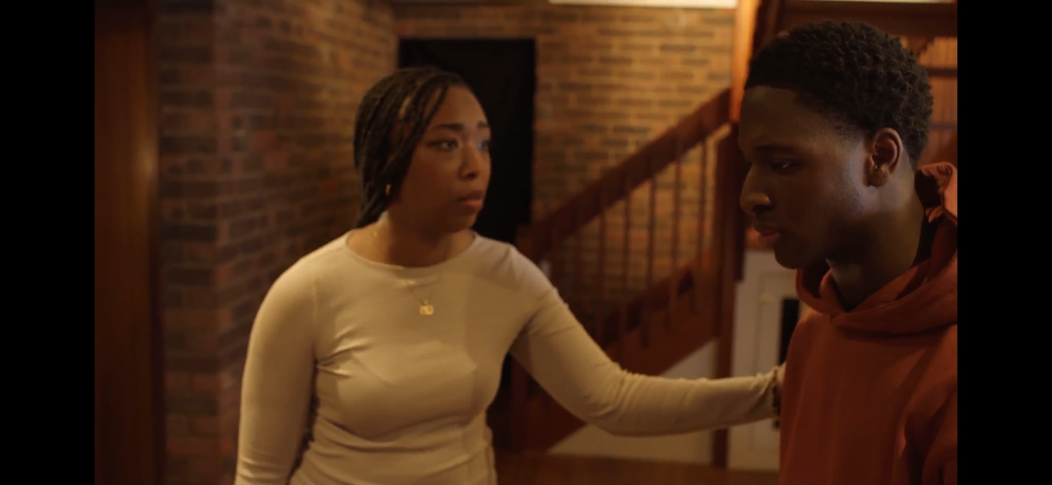 A young woman with braided hair wearing a light-coloured top stands beside a young man in an orange hoodie, who looks slightly downwards. They are in a room with exposed brick walls and a wooden staircase in the background.