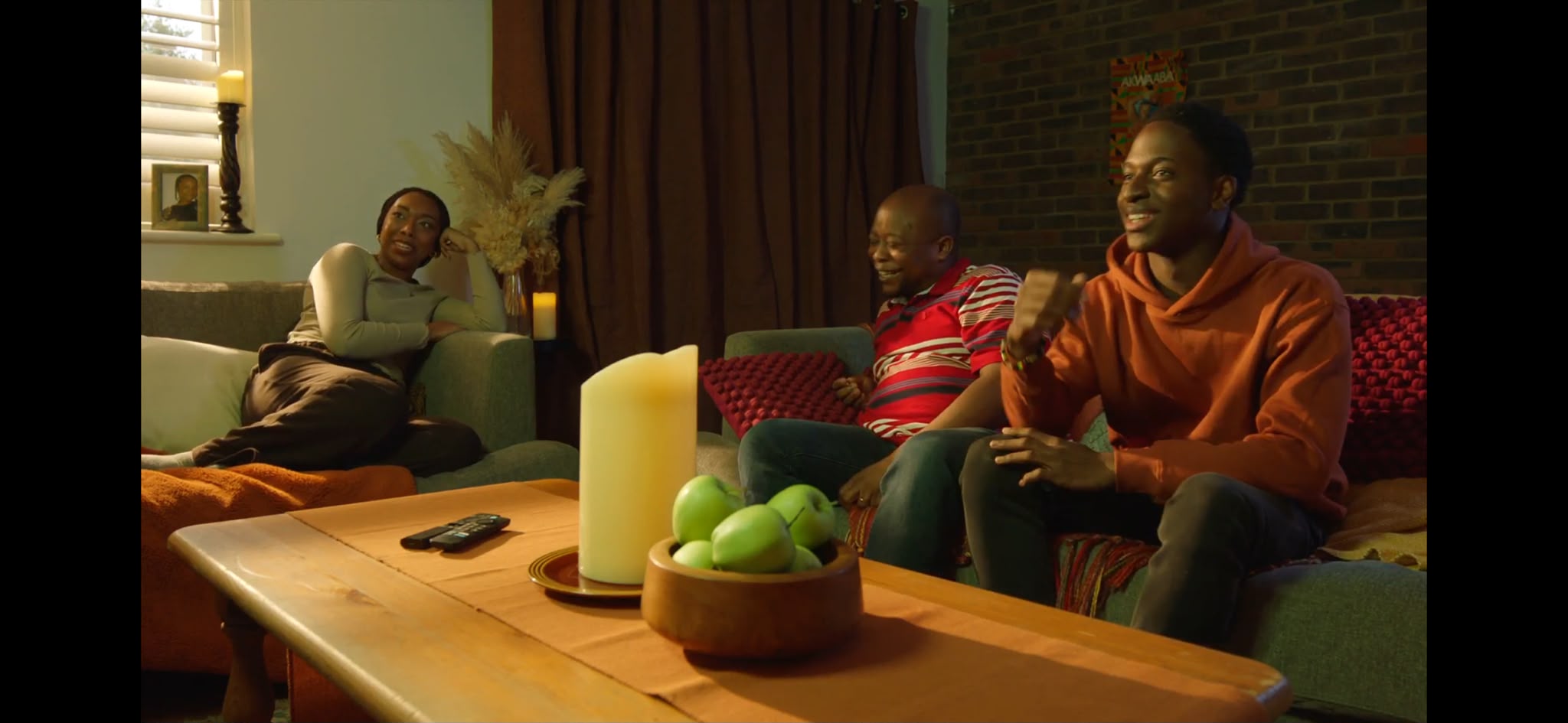 Three people are seated in a living room. A woman in a white top lounges on a green sofa, while a man in a striped shirt sits beside a younger man in an orange hoodie. A wooden coffee table with a candle and a bowl of green apples is in front of them.