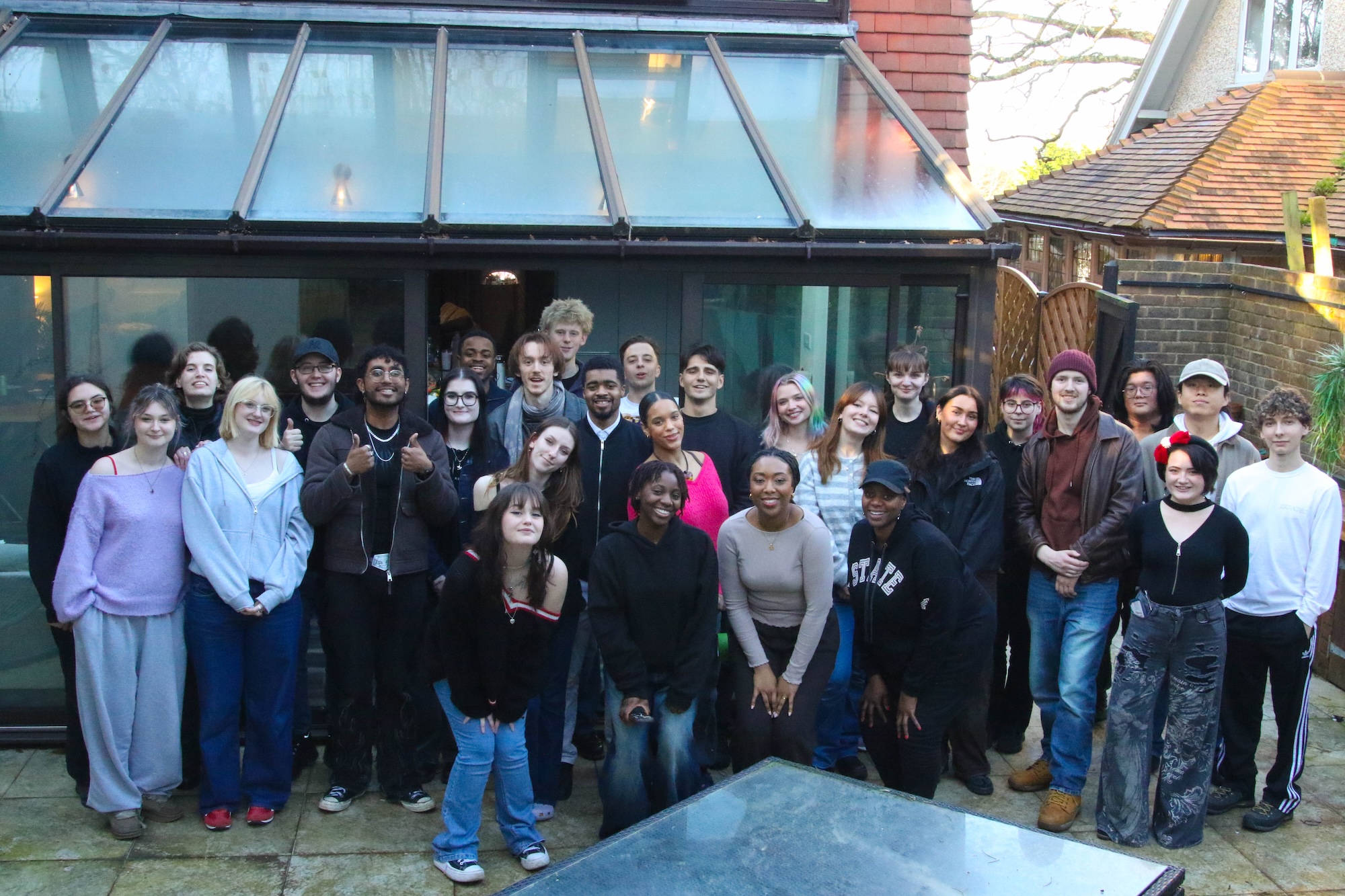 A film crew around 30 young adults stands outdoors in front of a glass-walled building. They are arranged in a semi-circle, with a mix of clothing styles and casual poses, and the background features a sloped roof and greenery.