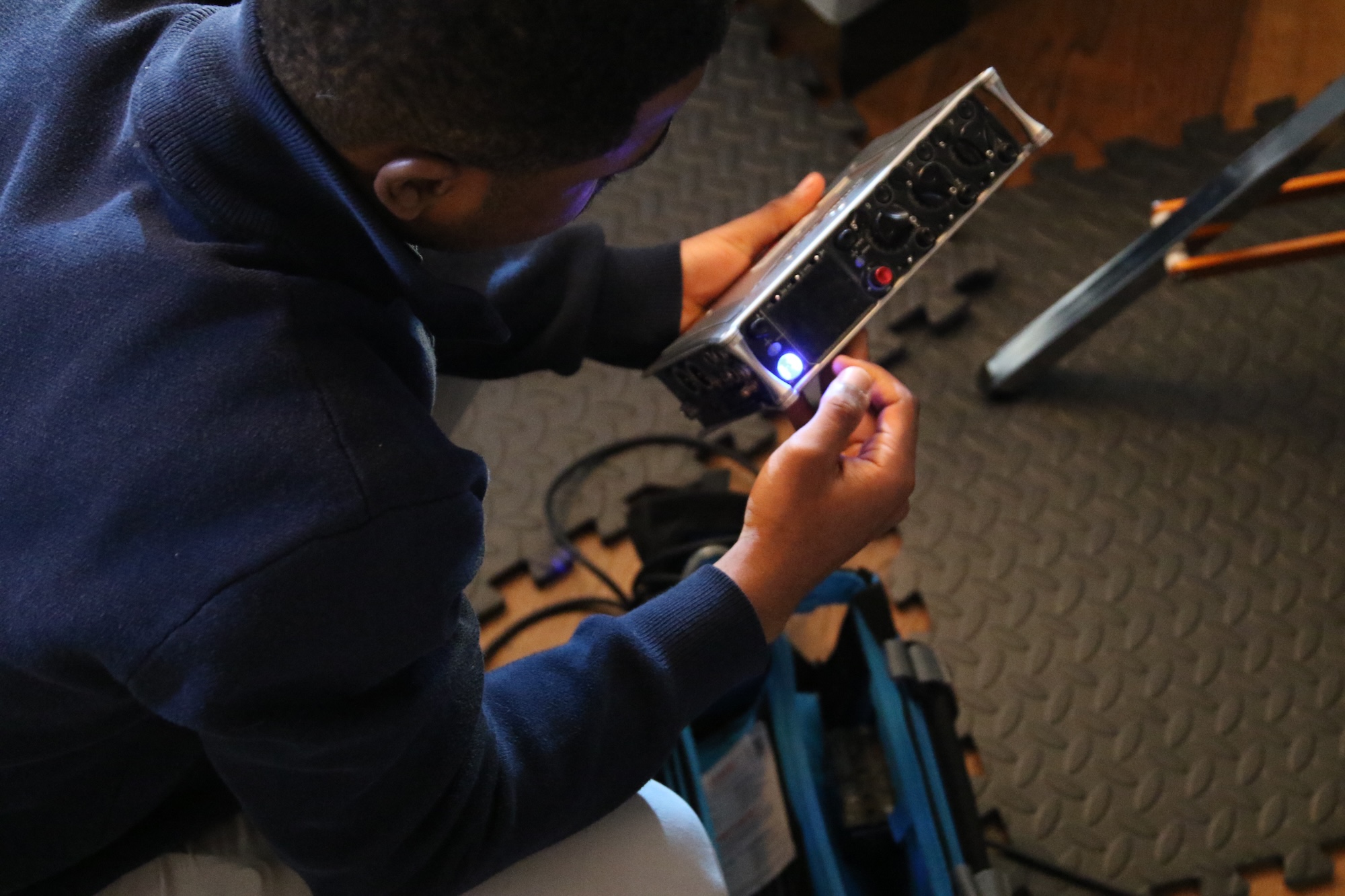 A young man in a navy blue sweater is seated on the floor, examining a silver equipment device with multiple buttons and a blue indicator light. Cables and a bag are visible nearby on a textured surface.