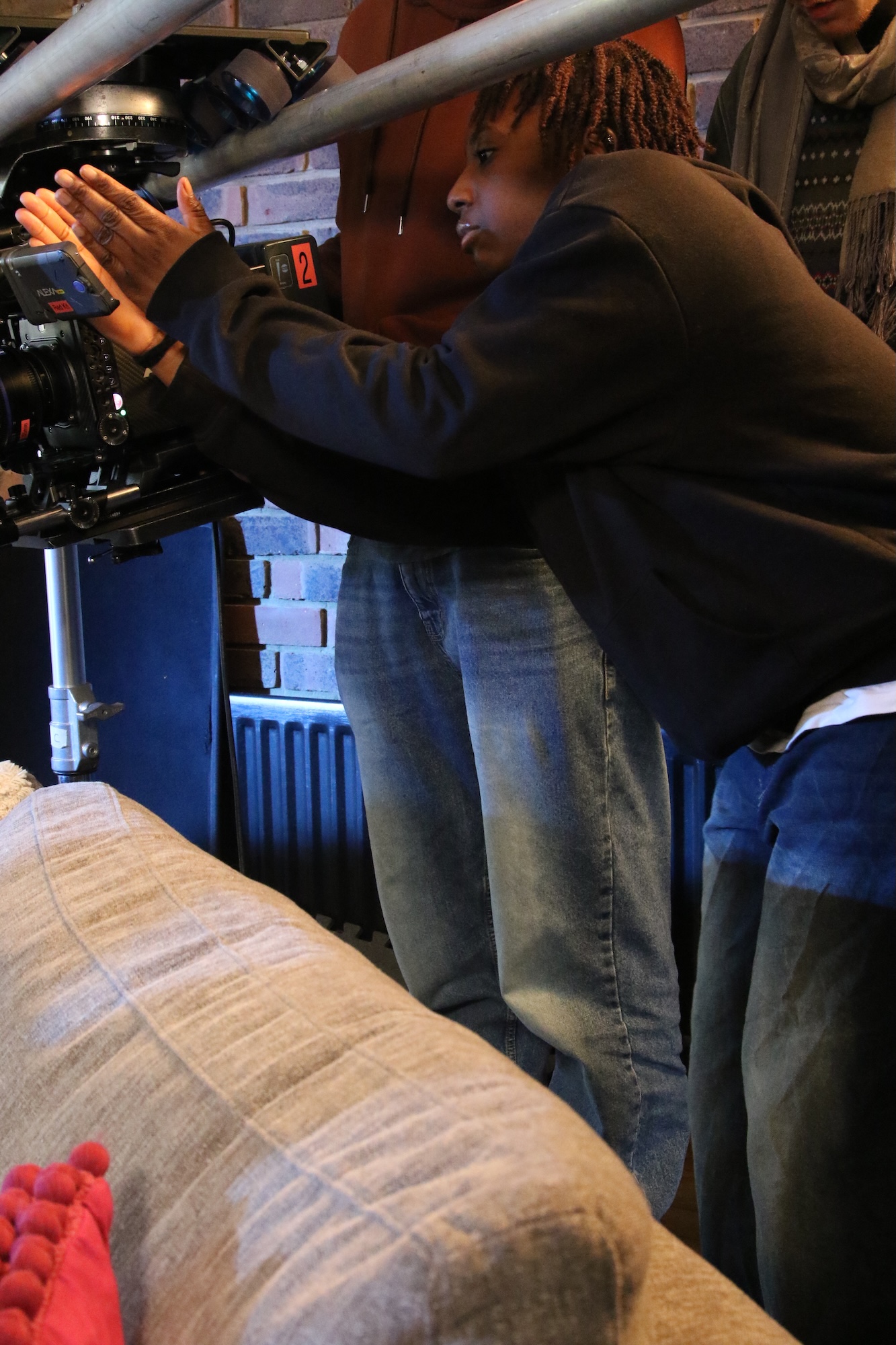 A person with dreadlocks operates a film camera on a tripod while leaning forward. In the background, part of a blue radiator and other crew members are visible, with a grey sofa in the foreground.
