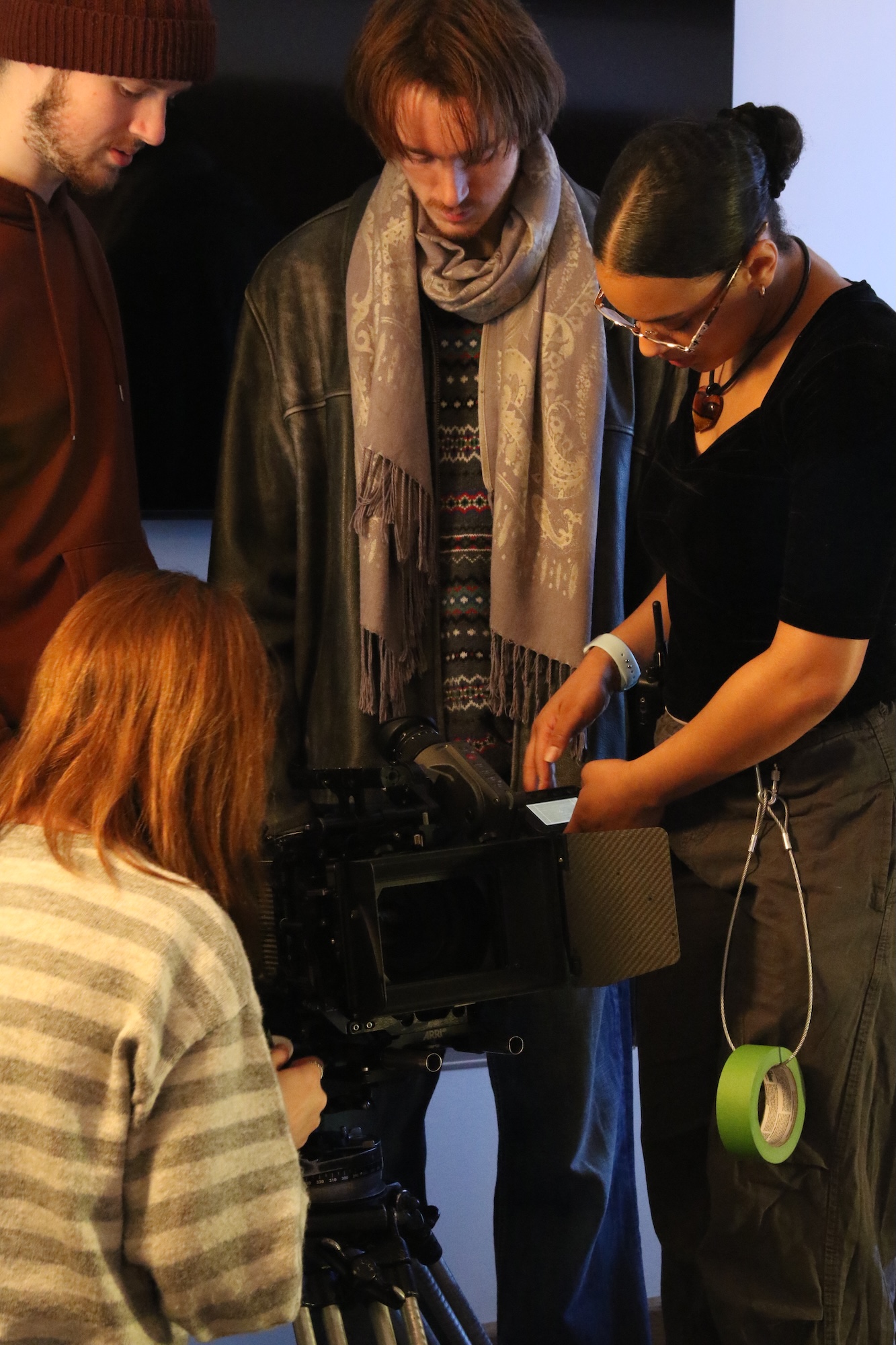 A group of four individuals interact around a film camera. A woman with glasses and a black shirt examines the camera, while another person with a scarf looks on. Two individuals, one in a brown sweater and another in a dark coat, observe the setup.