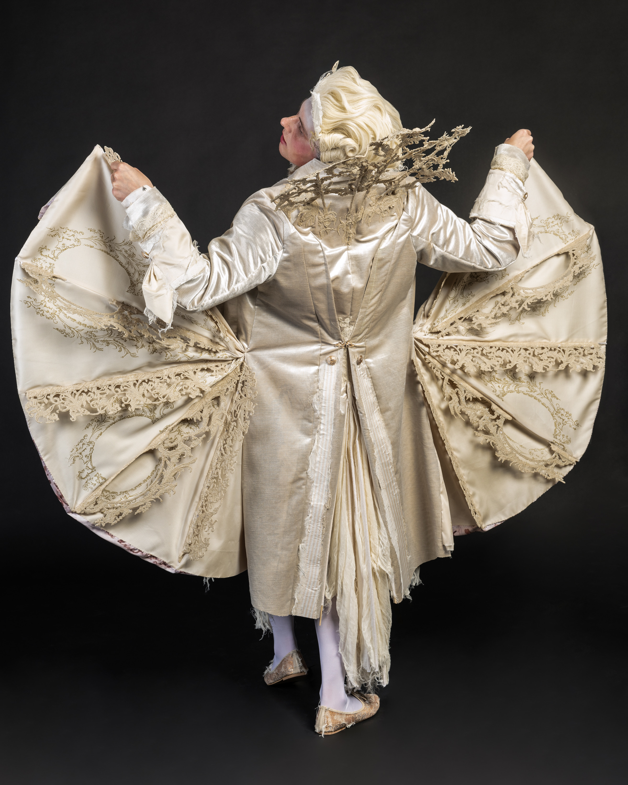 Person in flowing cream-coloured robe with wide sleeves outstretched, ornate golden embroidery, blonde hair with decorative headpiece.