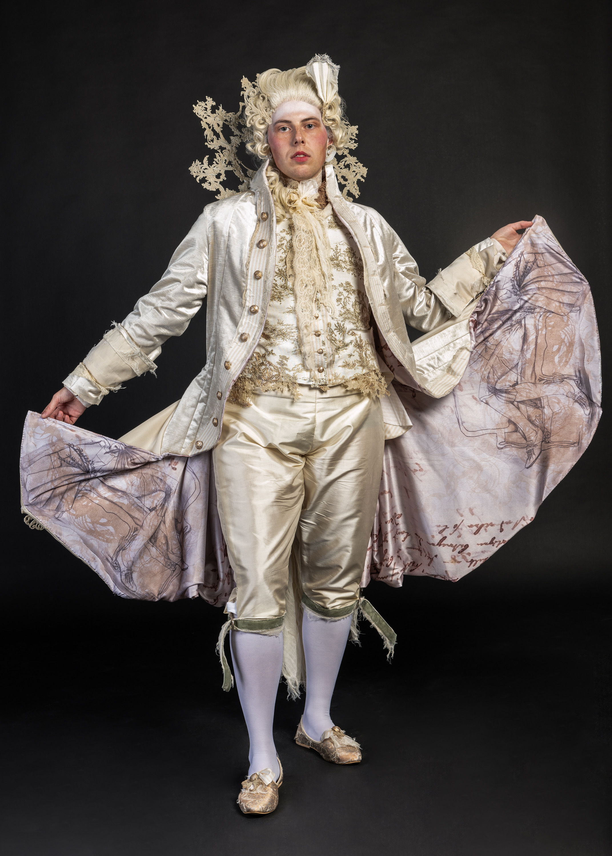 Performer in 18th-century costume with white powdered wig, cream silk coat and breeches, holding ornate cape with purple floral lining.