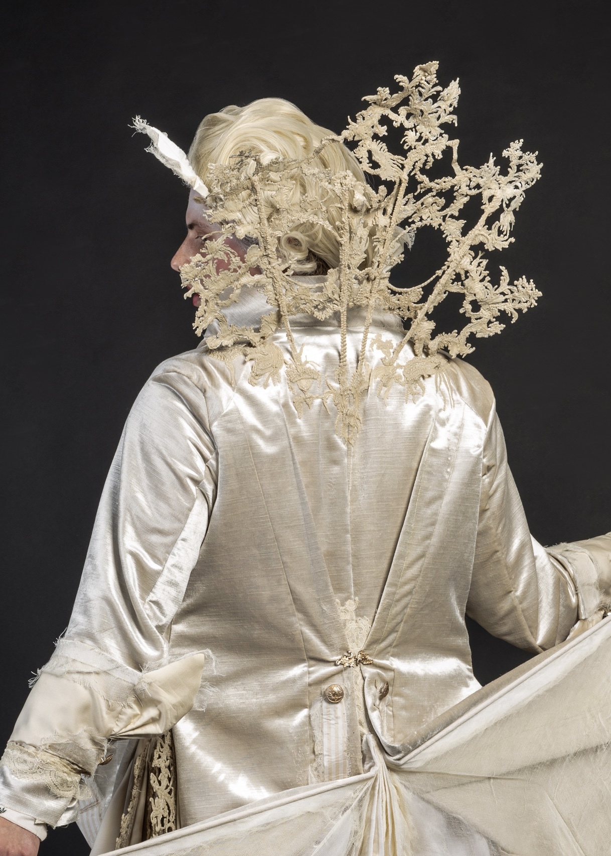 Person in cream silk garment with elaborate golden branch-like headpiece covering face, seated against dark background.