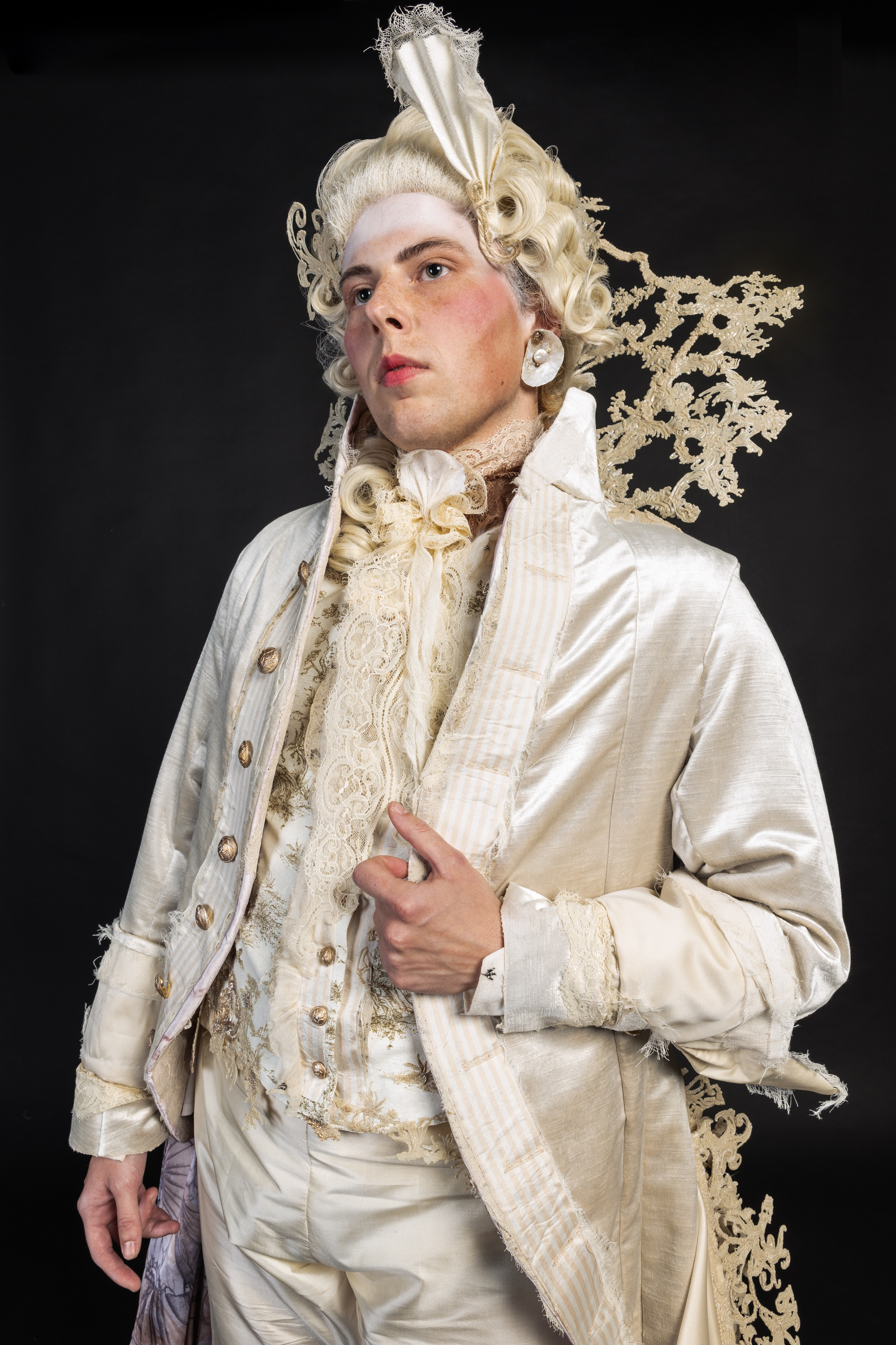 Actor in elaborate 18th-century costume with white powdered wig, cream silk coat, ornate lace cravat, and decorative fan against black background.