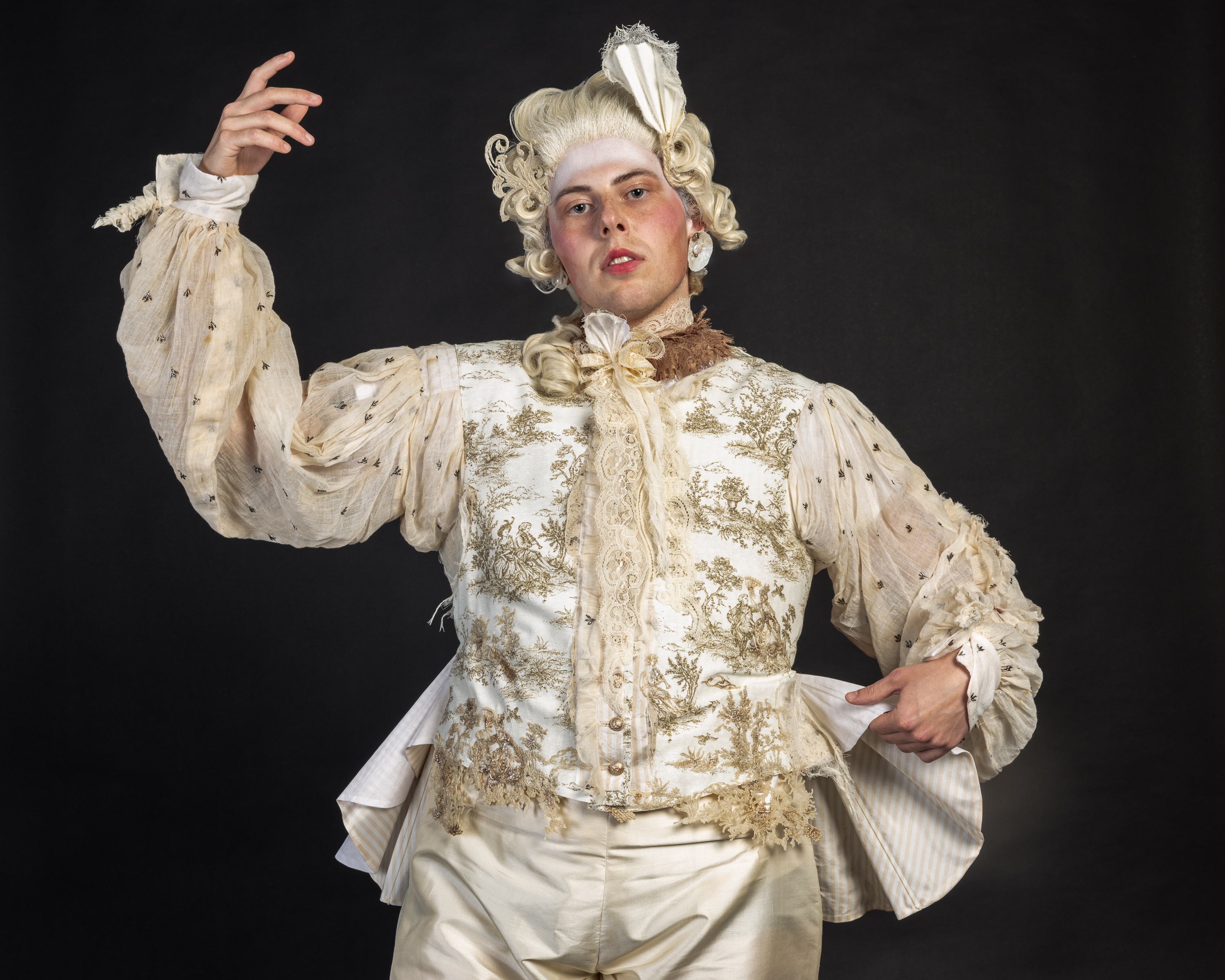 Person in elaborate white and gold period costume with powdered wig, ornate embroidered jacket, and ruffled collar against black background.