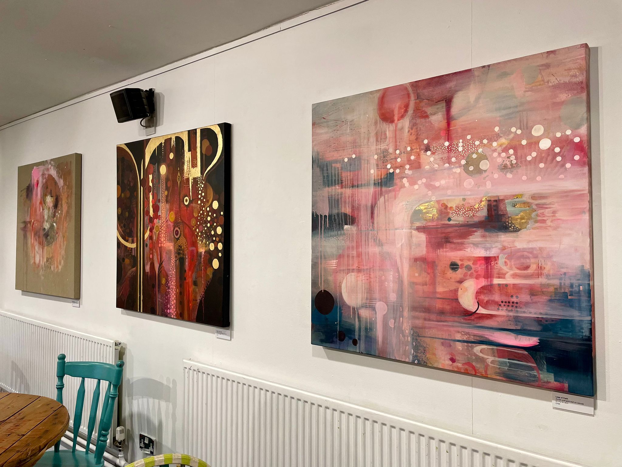 Three large abstract paintings on white wall: left shows muted tones, centre features dark background with golden swirls and white dots, right displays pink and blue layers with white circular elements.