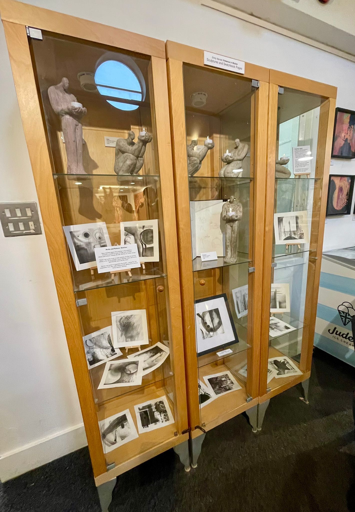 Wooden display cabinet with glass doors containing sculptures, photographs, and documents on multiple shelves in museum setting.
