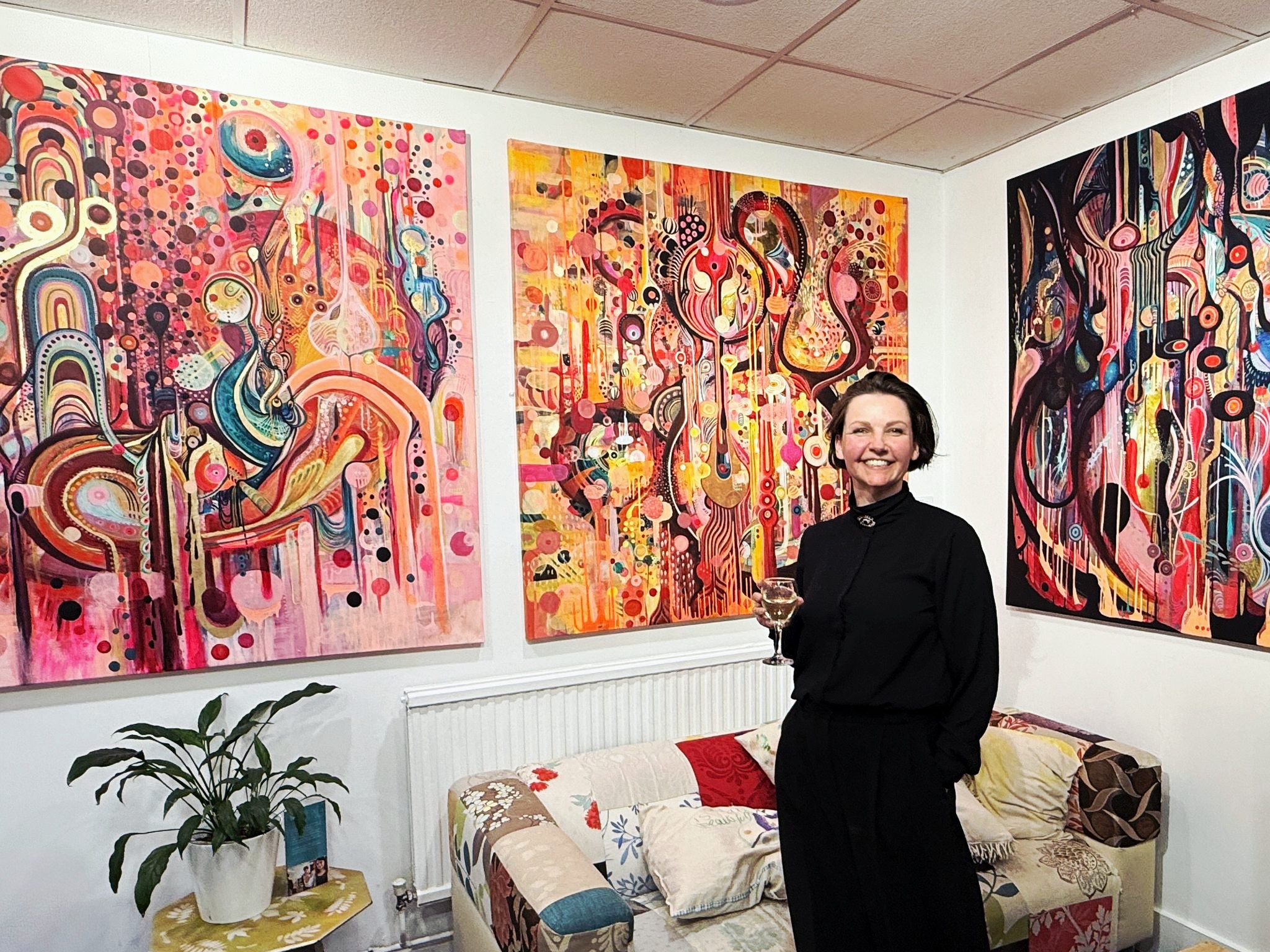 Woman in black dress stands in white room with three large colourful abstract paintings featuring swirling patterns, circles, and organic shapes in vibrant hues.