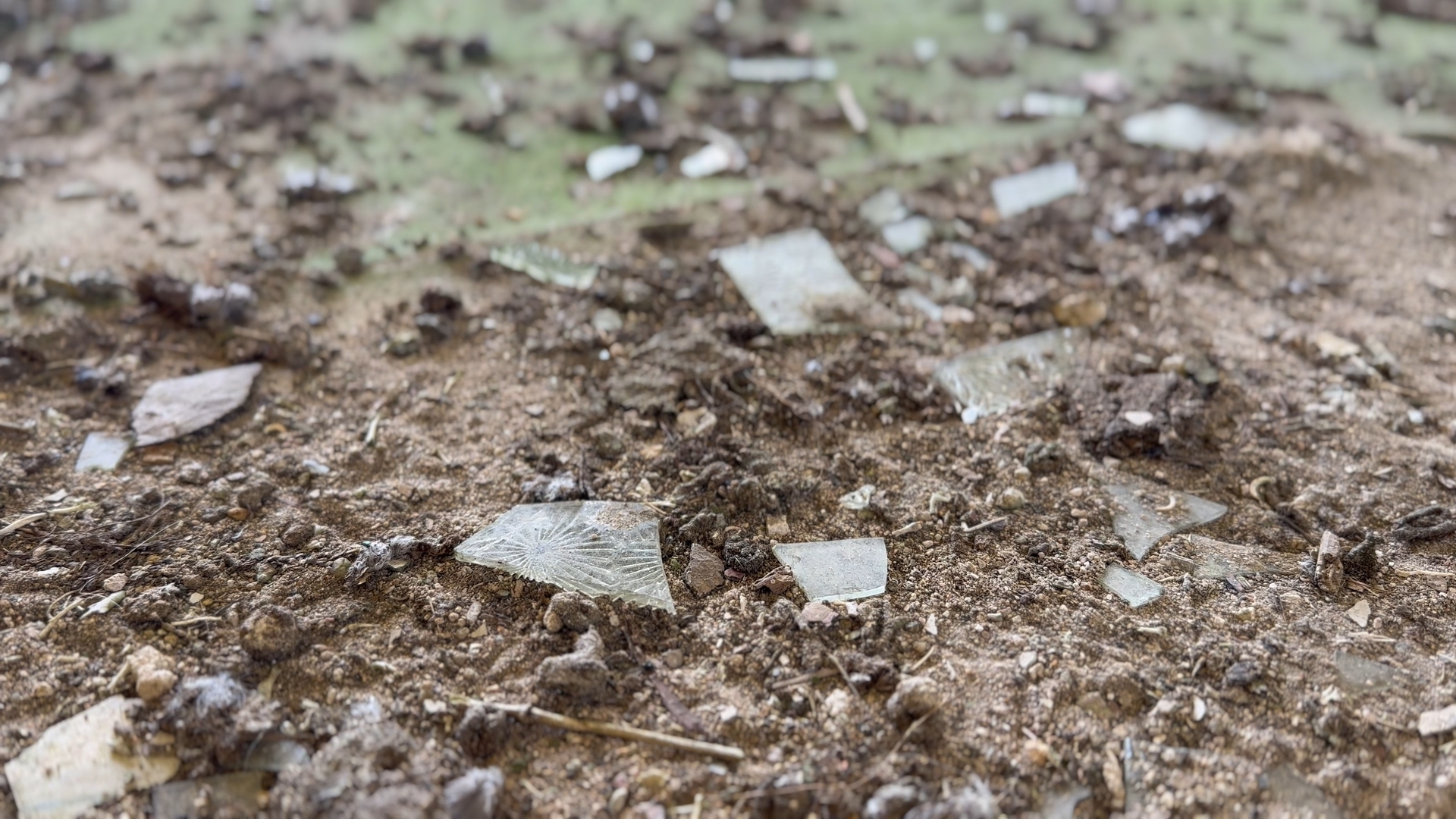 Broken glass fragments scattered across brown dirt and soil, with pieces reflecting light against earthy ground surface.