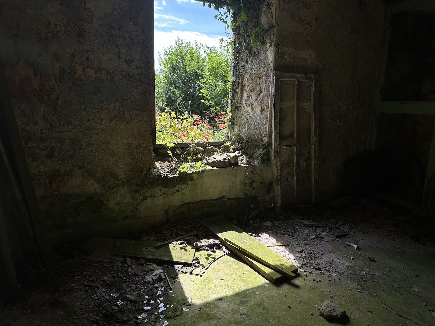 Interior of derelict room with crumbling walls, debris on floor, broken window frame revealing green trees and blue sky outside.