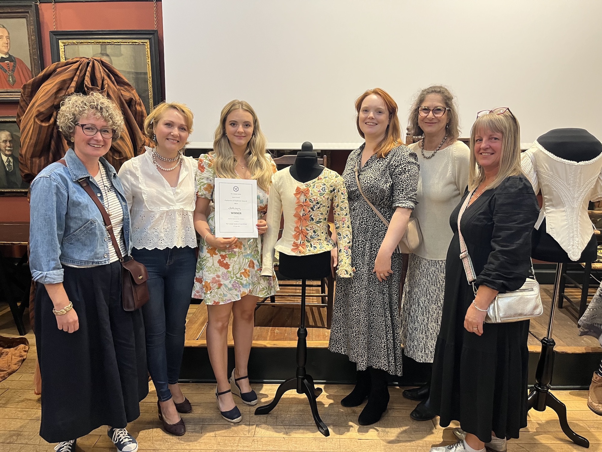 BA (Hons) Costume graduate Beth Lacey and members of the Costume course team posing for a photo at the Patterns of Fashion Awards 2023. The winning garment, an embroidered jacket, is displayed on a mannequin in the centre.