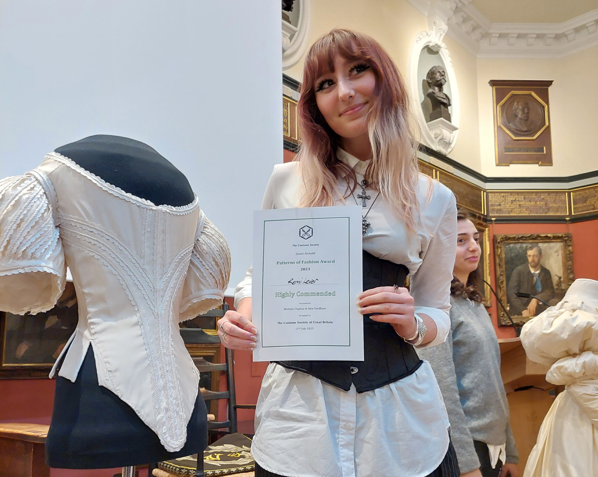 BA (Hons) Costume graduate Remi Gene Lever posing with a certificate next to a 17th century bodice, on display on a mannequin, at the 2023 Patterns of Fashion Awards.