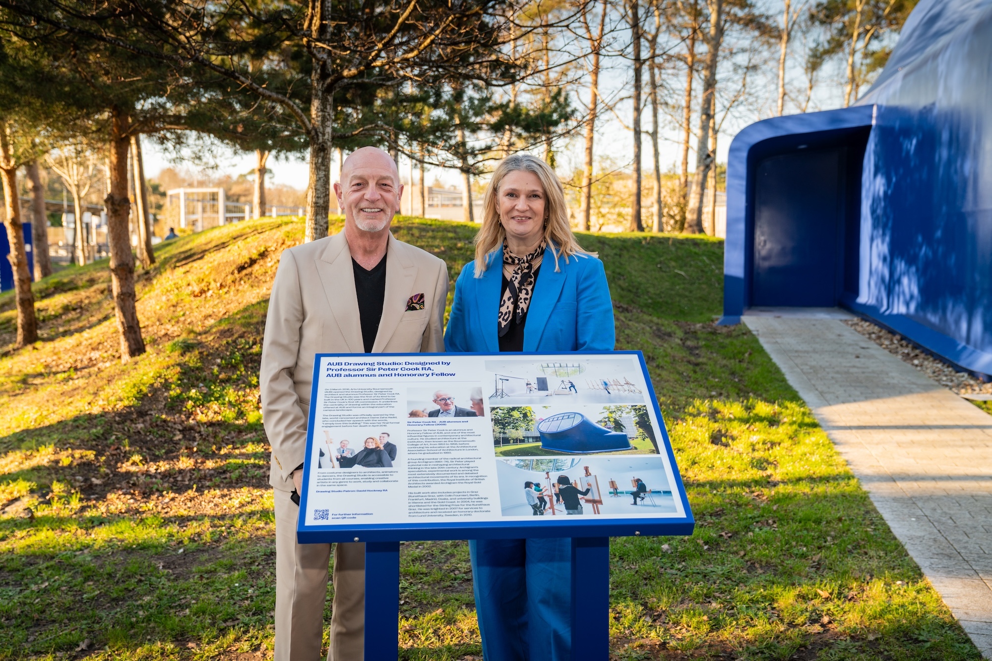 Two people standing behind blue information board outdoors, man in beige suit, woman in bright blue jacket, trees and blue structure behind.