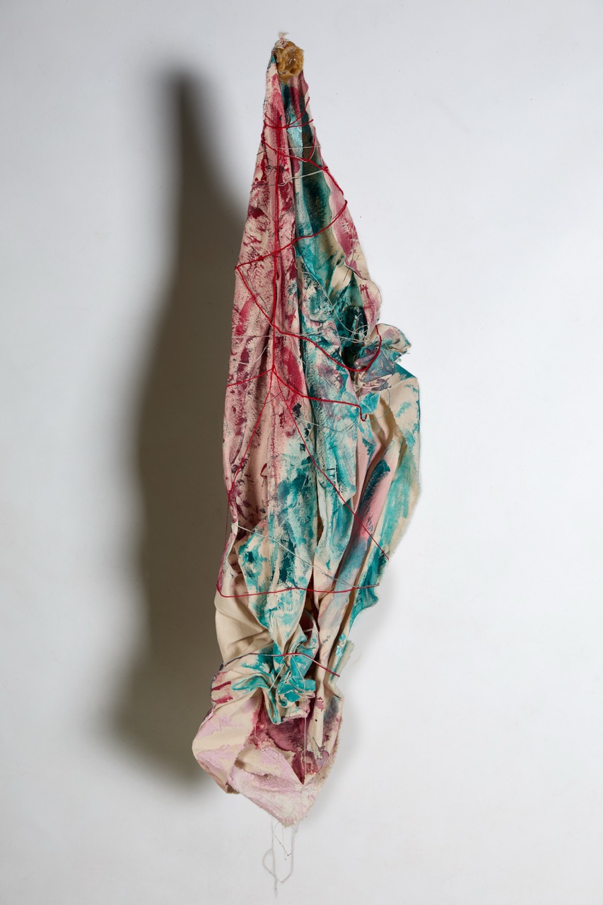 A large, crumpled piece of fabric featuring a mix of colours including pink, teal, and cream. The fabric is suspended from a hook, displaying abstract patterns and folds.