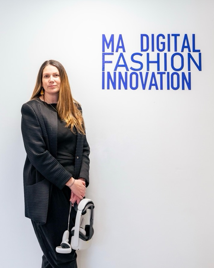 Penelope Norman stood in from of a white background holding white VR set, dressed in black, MA Digital Fashion Innovation in blue on the wall to the right of her.