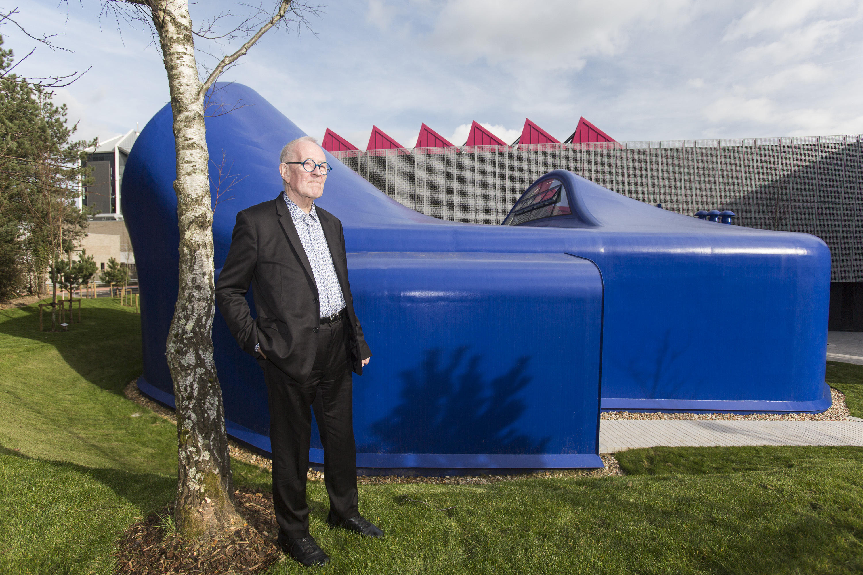 Man in dark suit standing beside large blue aircraft fuselage display with red triangular spikes on building behind