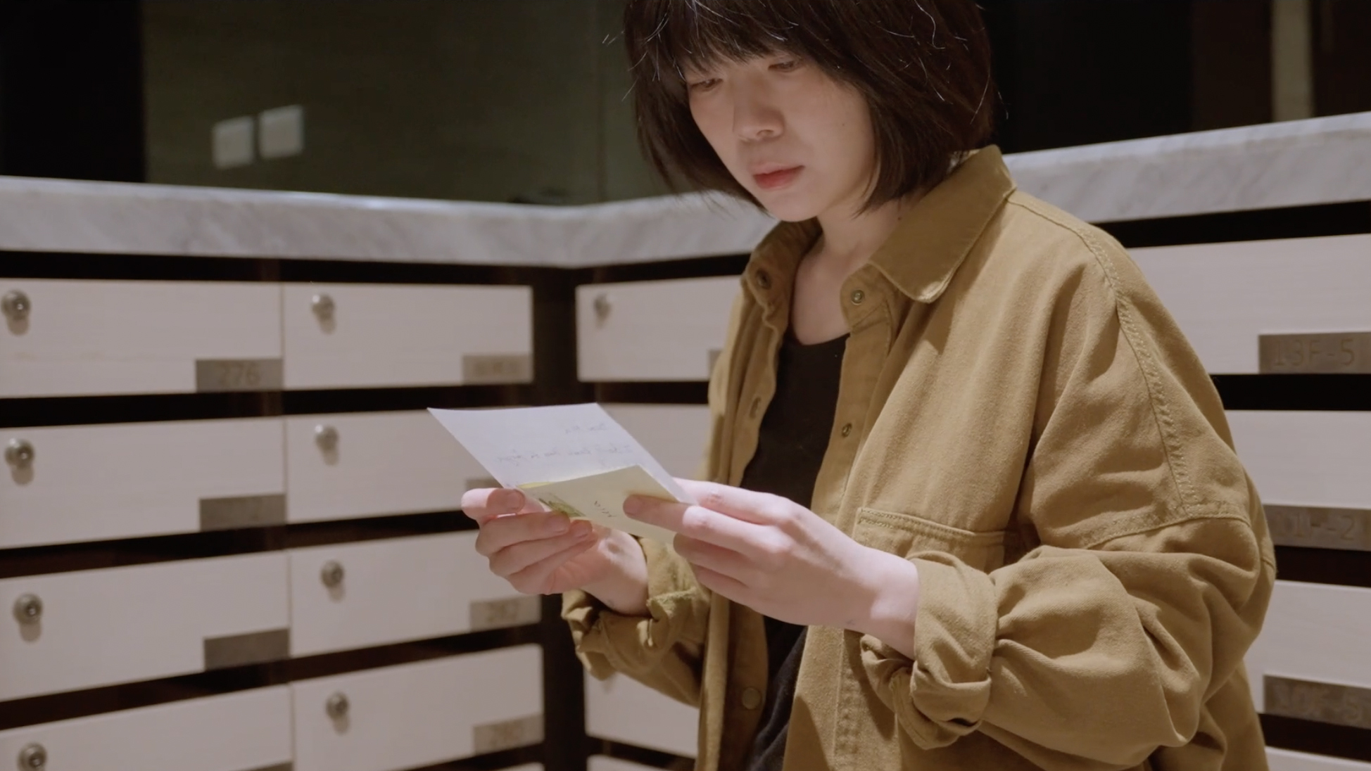 Person with short brown hair wearing beige jacket reading paper whilst standing by white railing with filing cabinets behind.